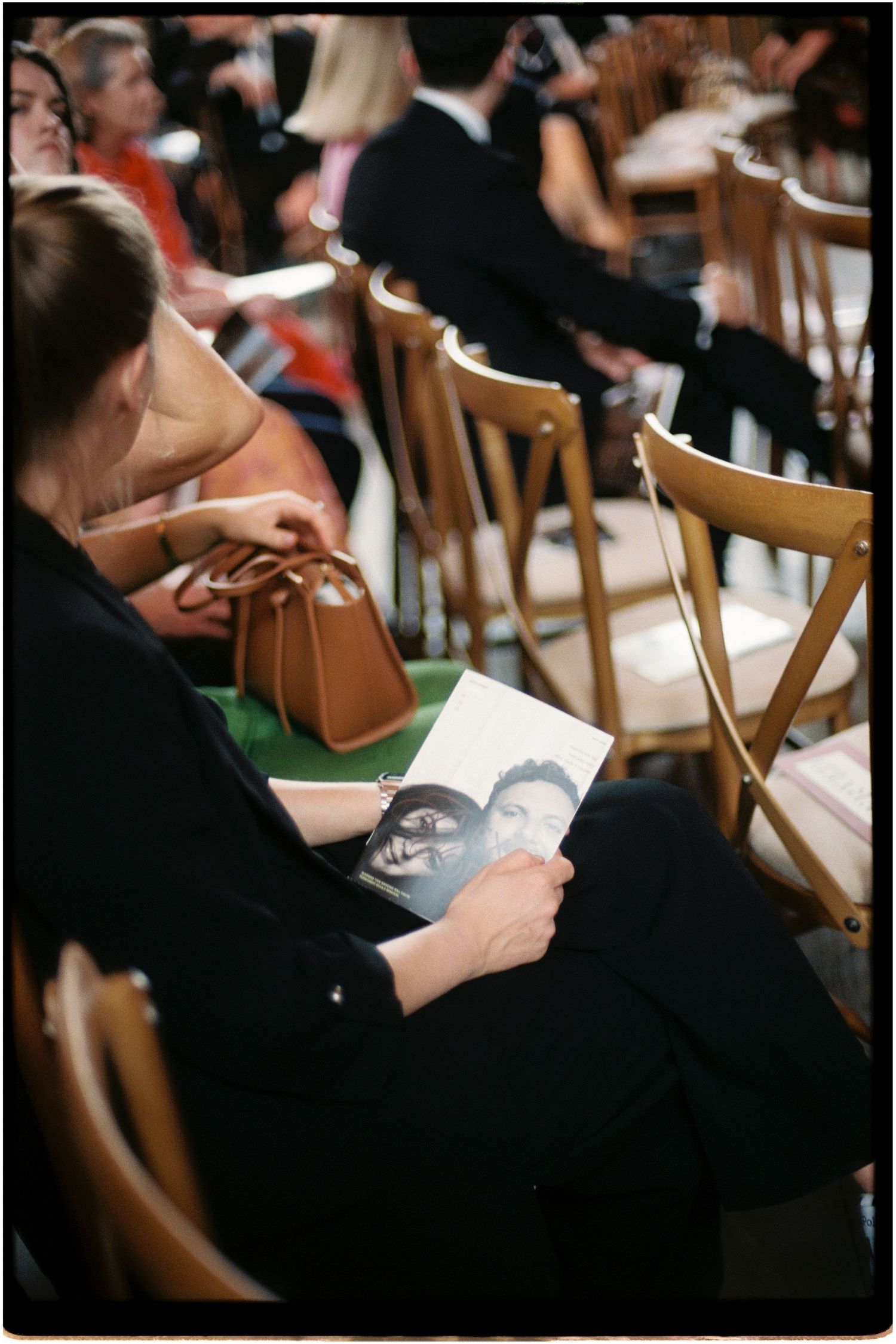 Wedding guest in black outfit reading a program with couple's photo at a formal ceremony.