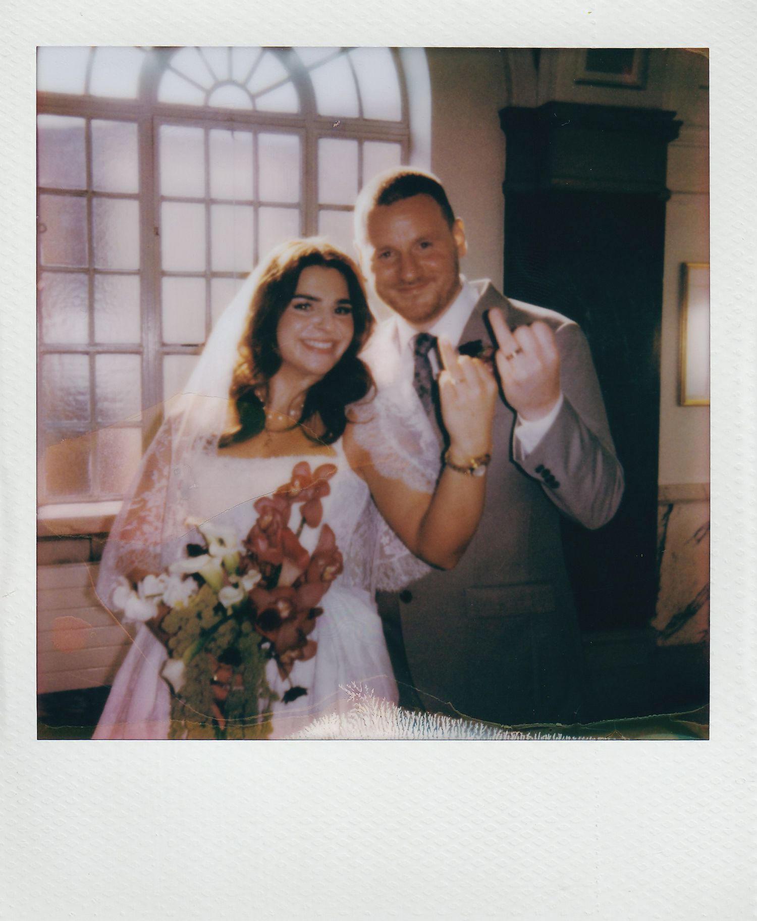 Smiling newlyweds posing for vintage Polaroid photo inside wedding venue with floral bouquet.