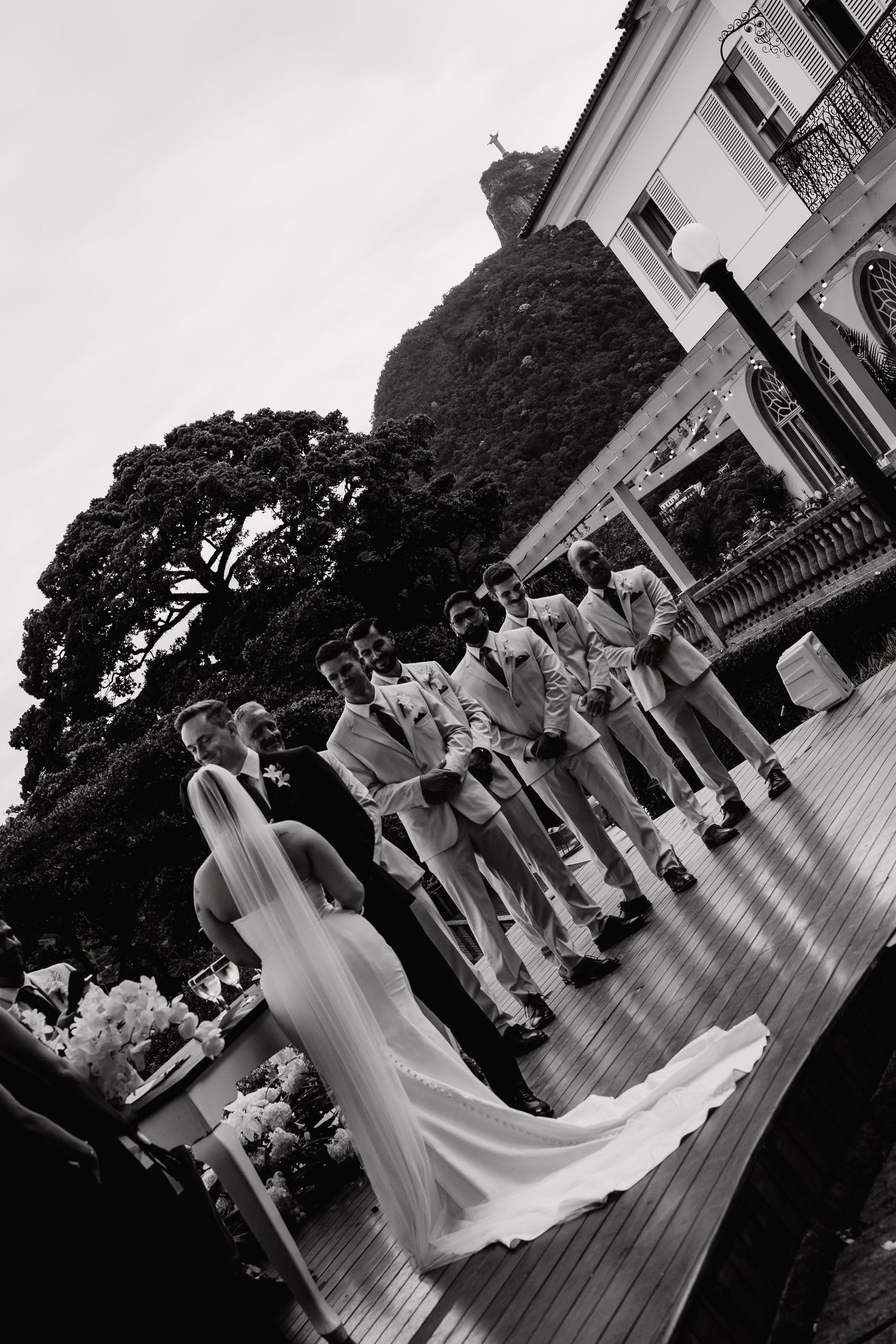 Bride and groom stand before wedding party on outdoor deck with lush trees in black and white.