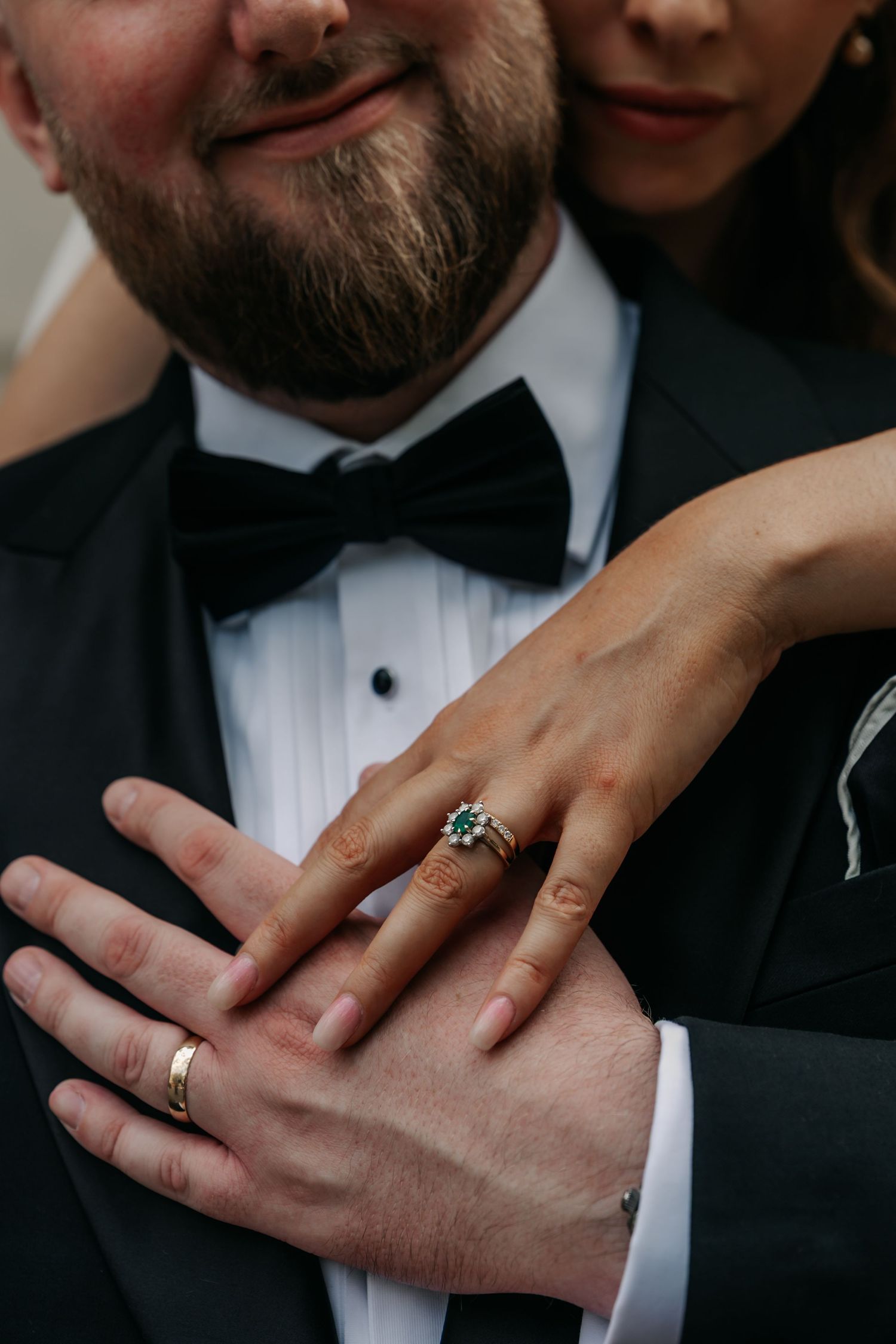 Close-up of bride and groom hands showing wedding rings, groom in black tuxedo with bow tie.