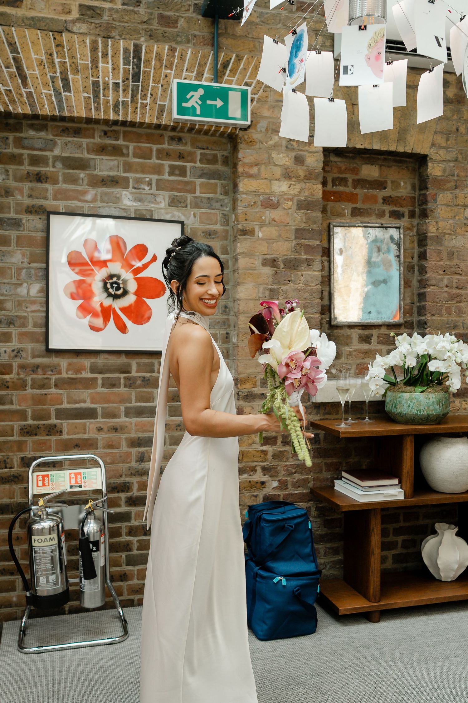 Bride in a sleek white halter gown holding a bouquet inside a stylish brick-walled wedding venue.
