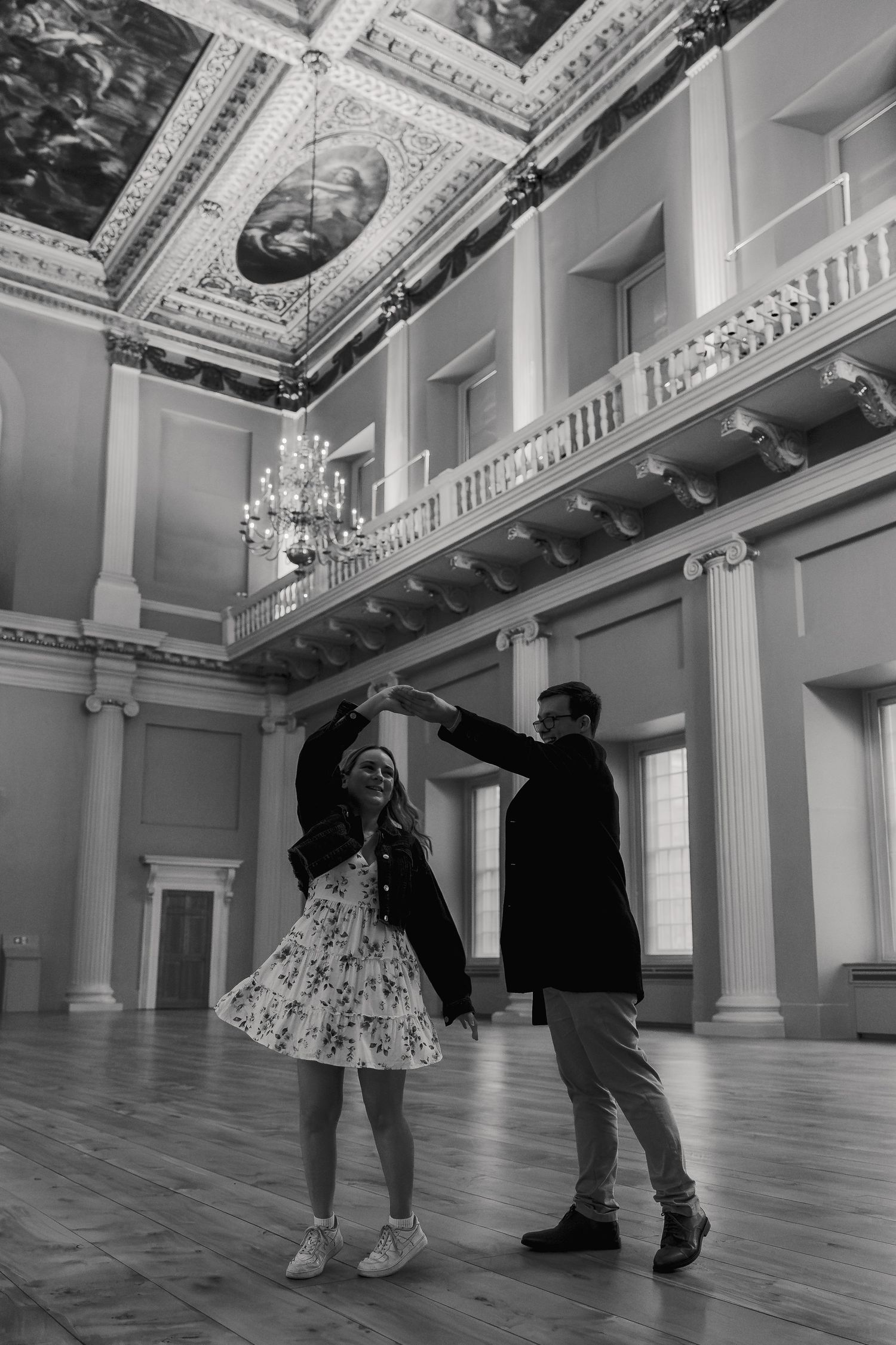 Black and white photo of couple dancing together in a stunning neoclassical ballroom with chandelier.