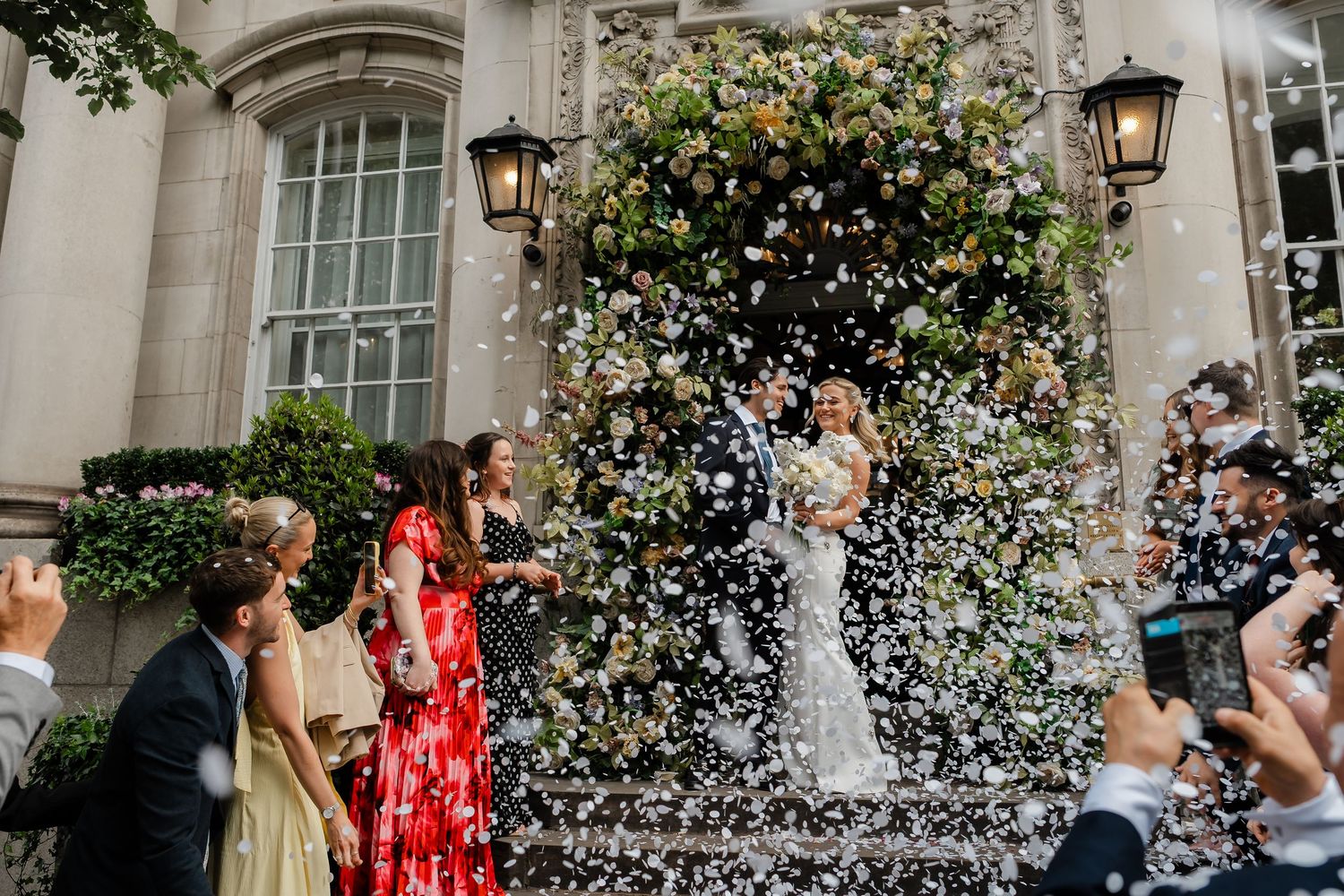 Guests shower newlyweds with confetti outside flower-adorned wedding venue entrance.