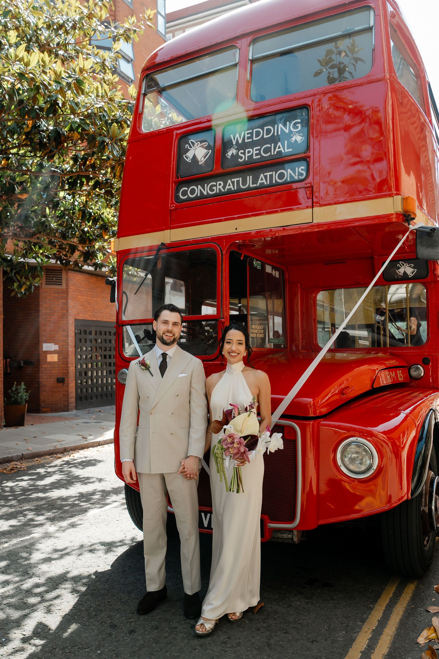 Bride and groom posing before a red London double-decker wedding special congratulations bus on a sunny street.