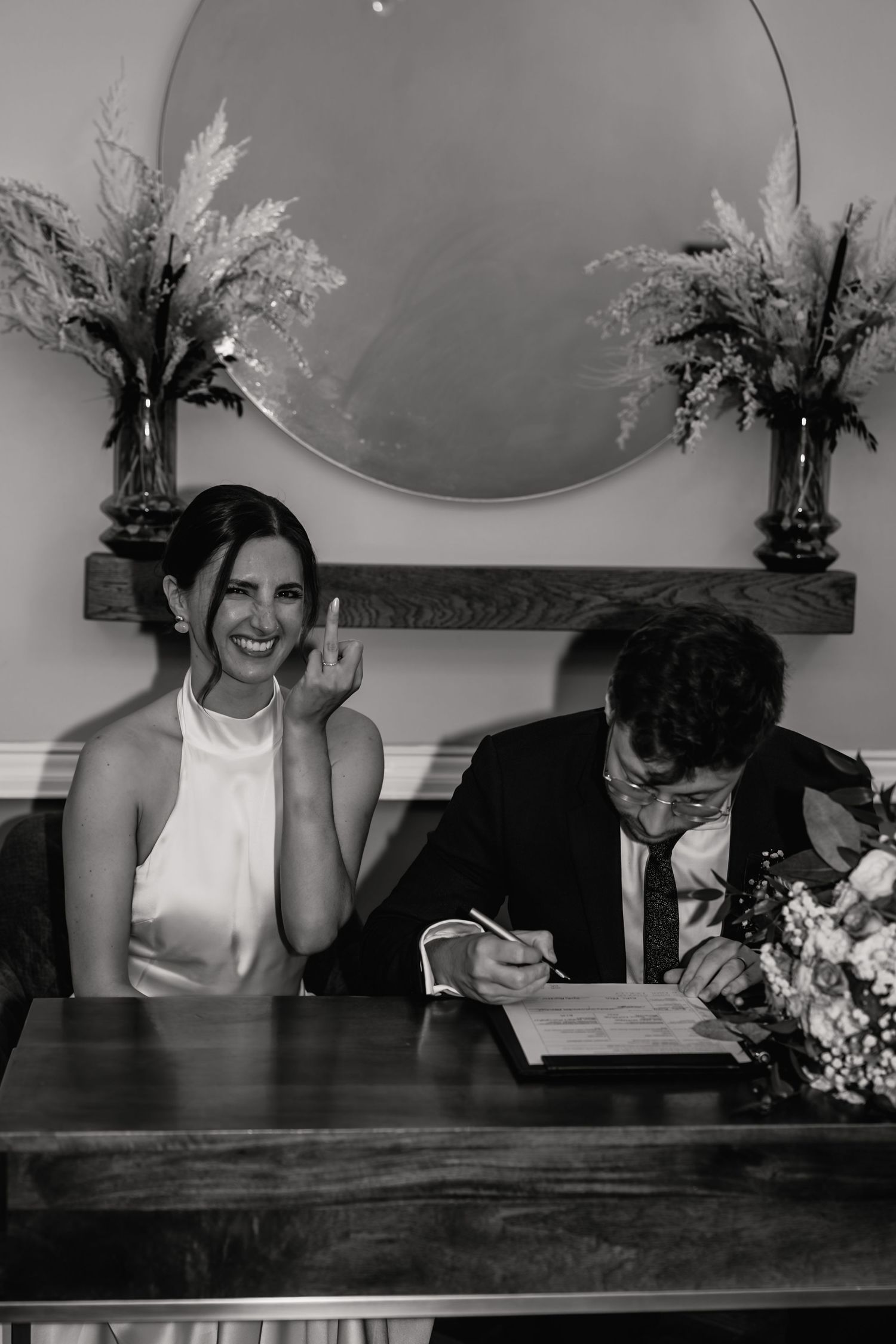 Black and white photo of a smiling bride in white dress and groom signing wedding documents at a wooden table.