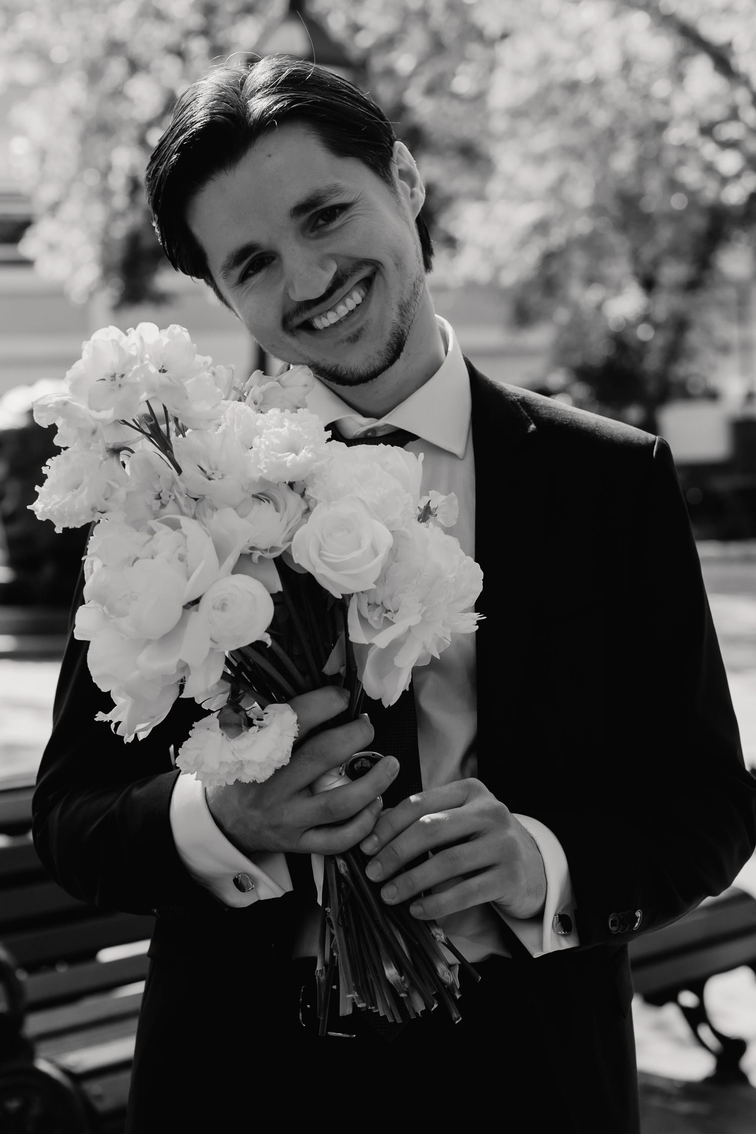Smiling groom in black suit holding large white floral bouquet outdoors in black and white photo.