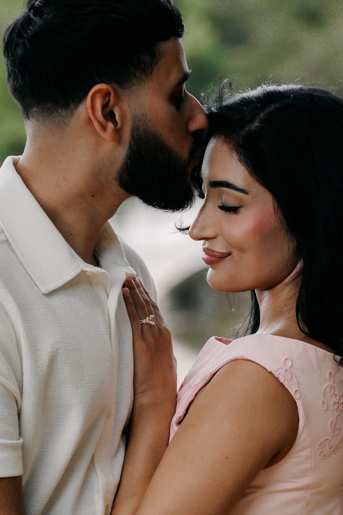 Couple sharing an intimate moment, man kissing woman's forehead as she smiles holding his chest showing engagement ring.