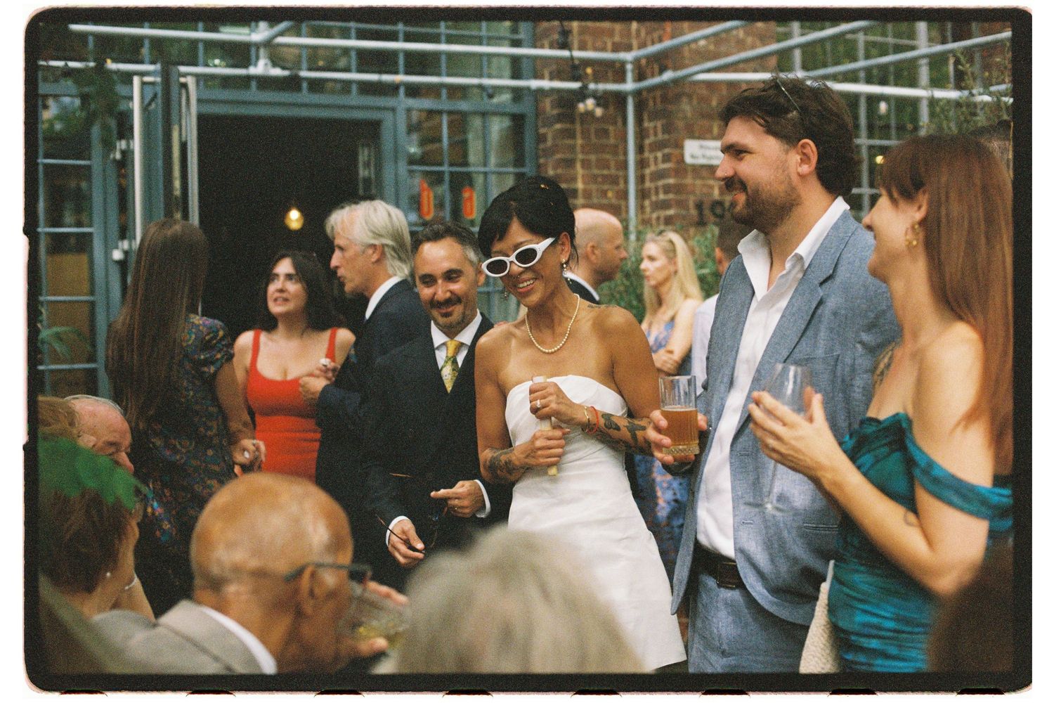 Bride in white dress with sunglasses mingles with guests at an outdoor wedding reception.