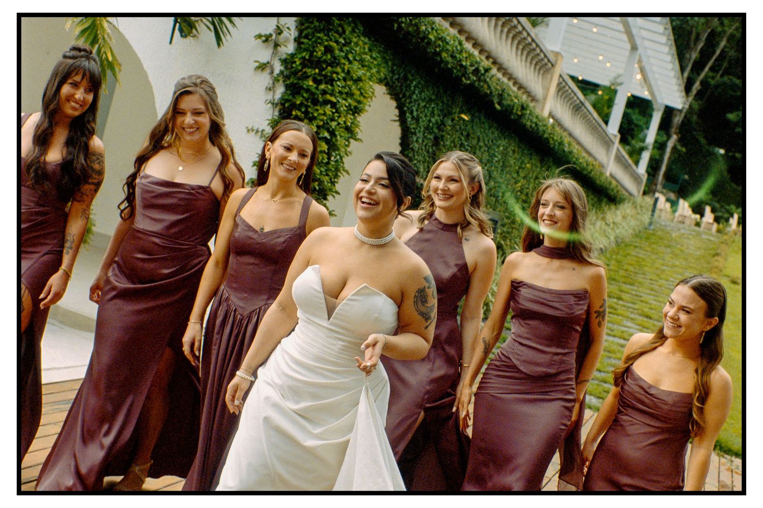 Bride smiling with six bridesmaids in dark burgundy dresses against an ivy-covered wall.