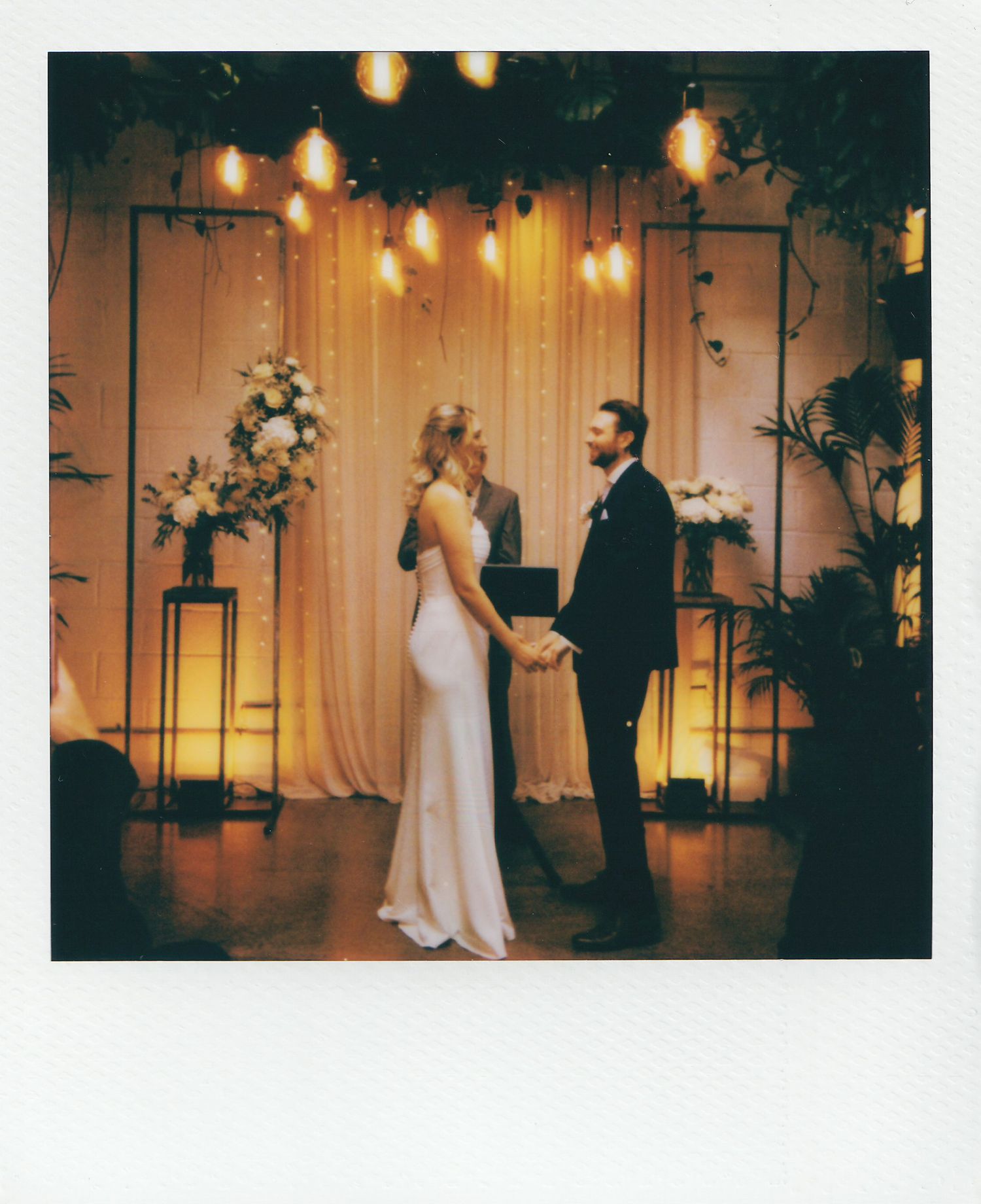 Couple exchanges vows at romantic indoor wedding ceremony with warm Edison bulb lighting and floral arrangements.