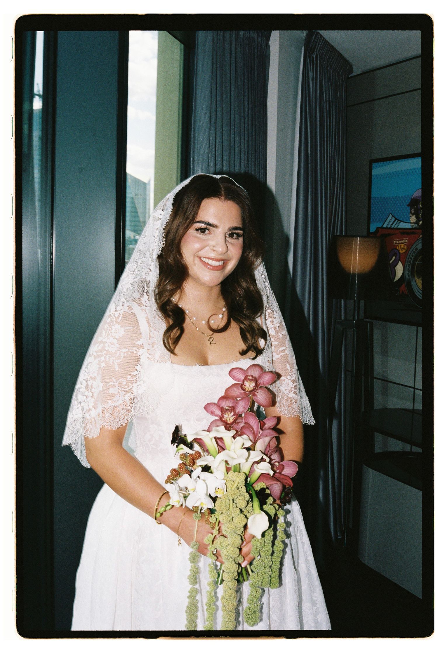 Smiling bride in white lace wedding dress and veil holding colorful orchid bouquet standing by window.