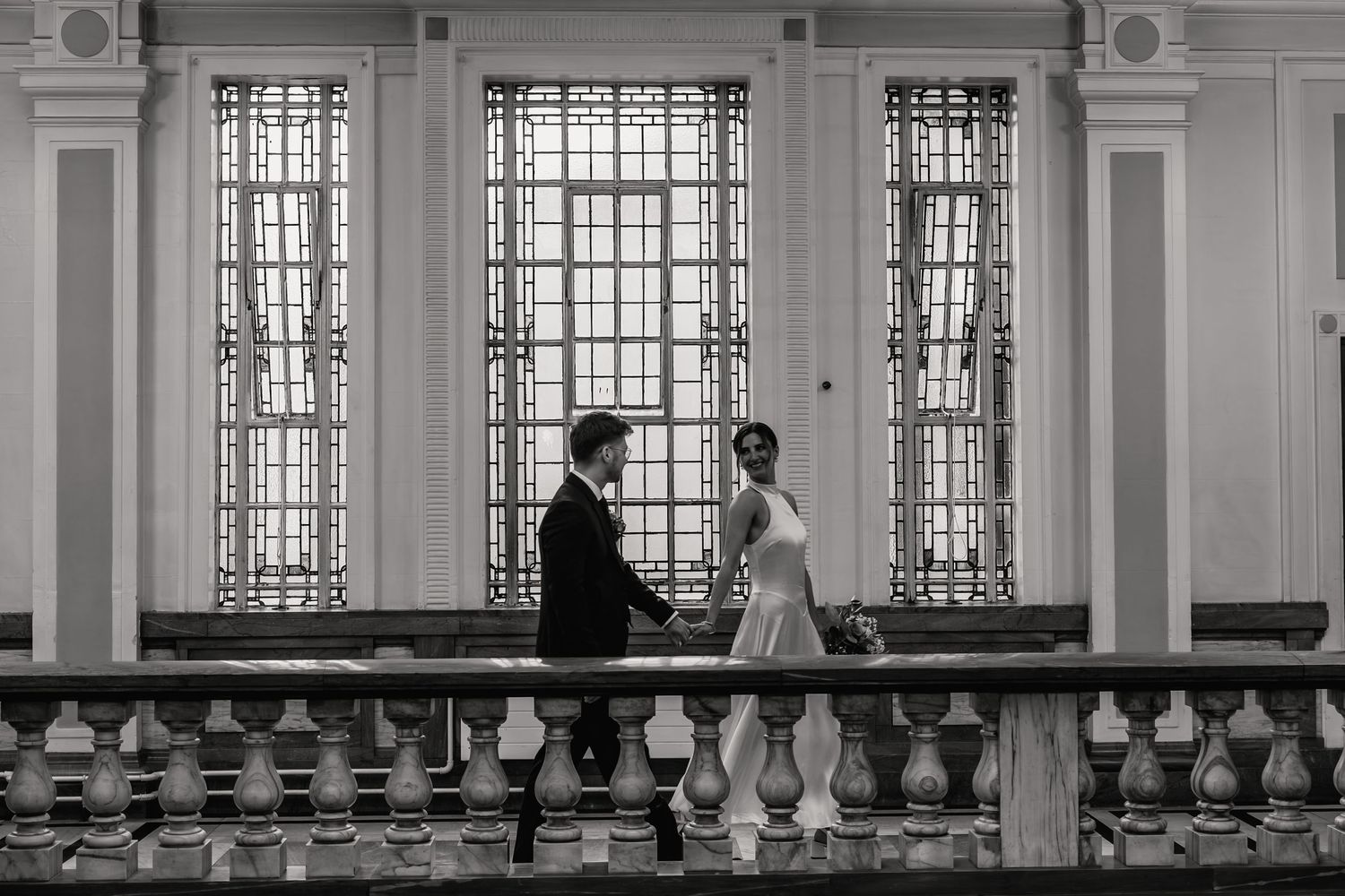 Black and white photo of a bride and groom walking hand in hand past elegant windows in a grand venue.