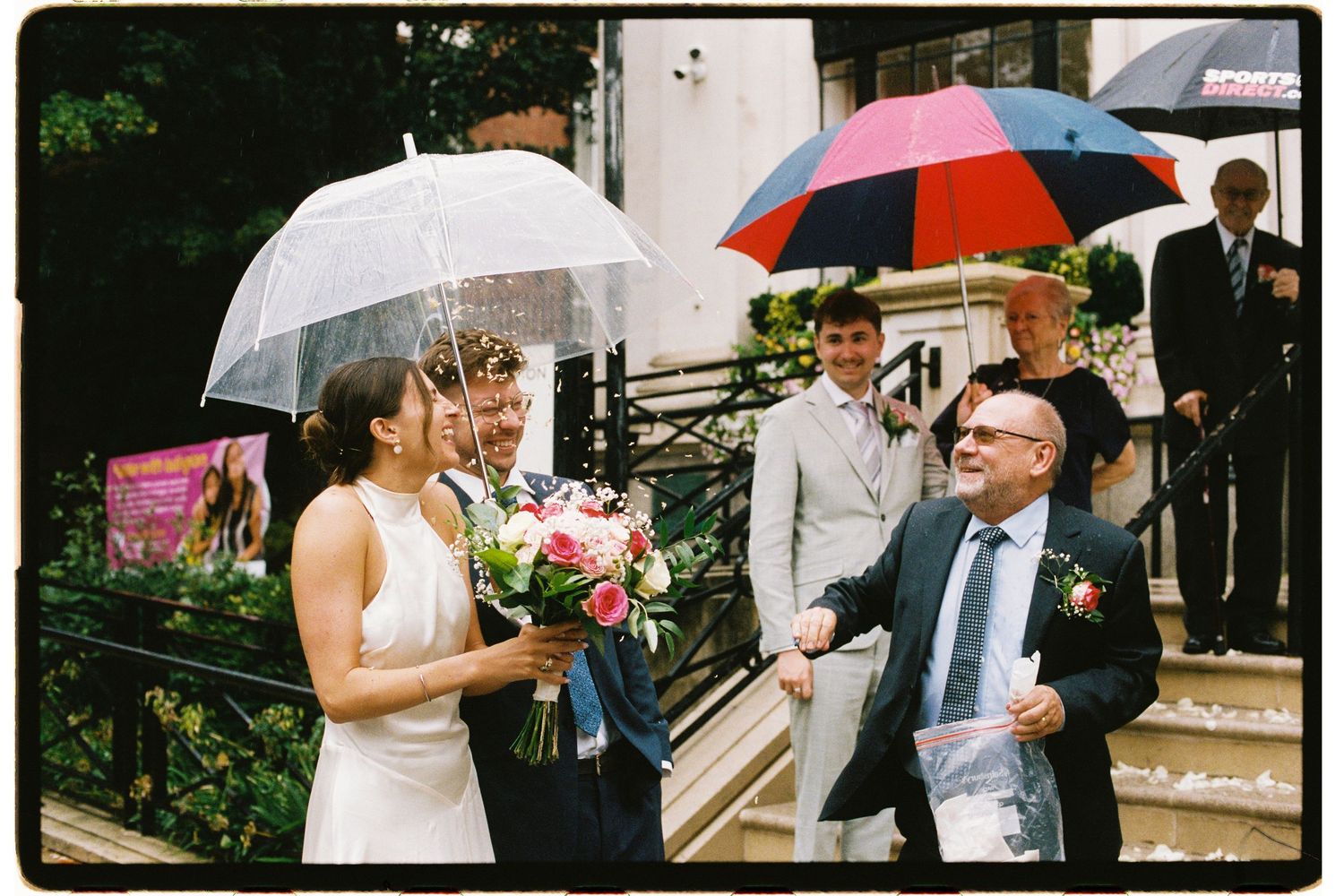 Bride and groom share a romantic moment under clear umbrellas on rainy wedding day outside venue.