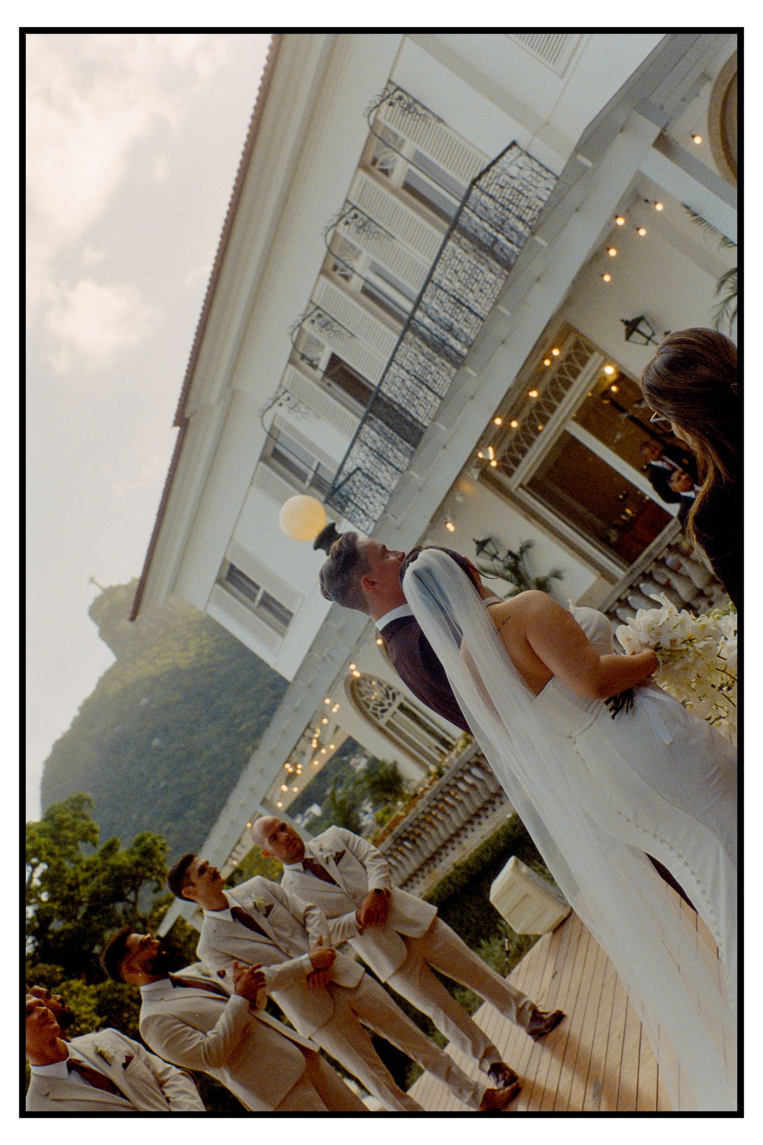 Wedding ceremony moment with couple kissing at altar, groomsmen in white suits, tropical venue.
