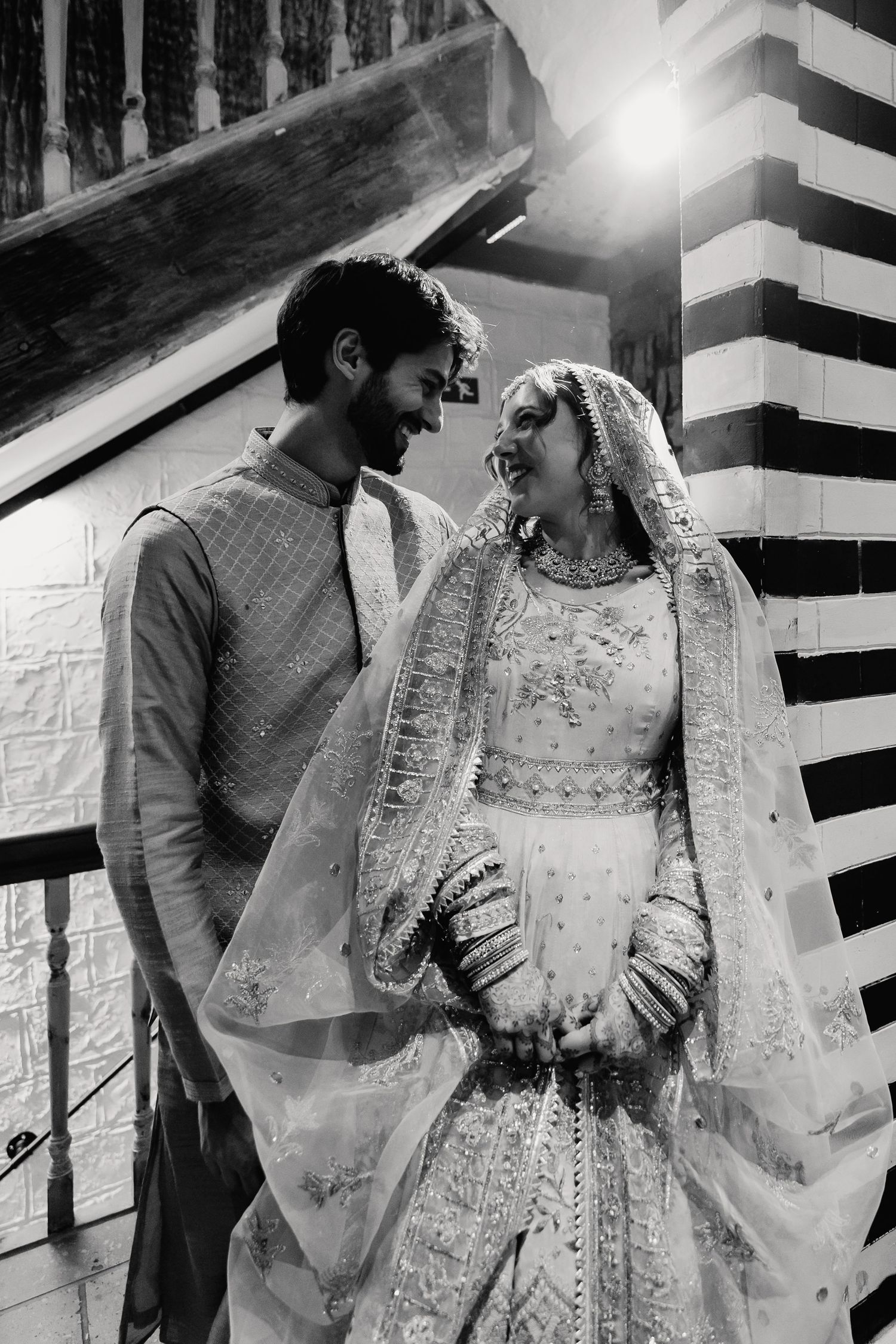Black and white portrait of South Asian bride and groom in traditional attire smiling at each other tenderly.
