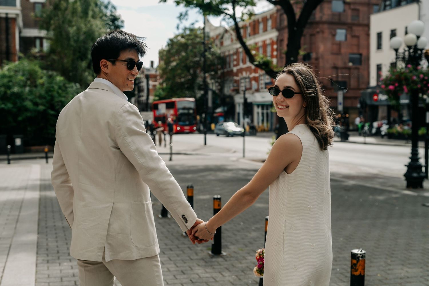 Newlywed couple holding hands and smiling on a London street in casual wedding attire.