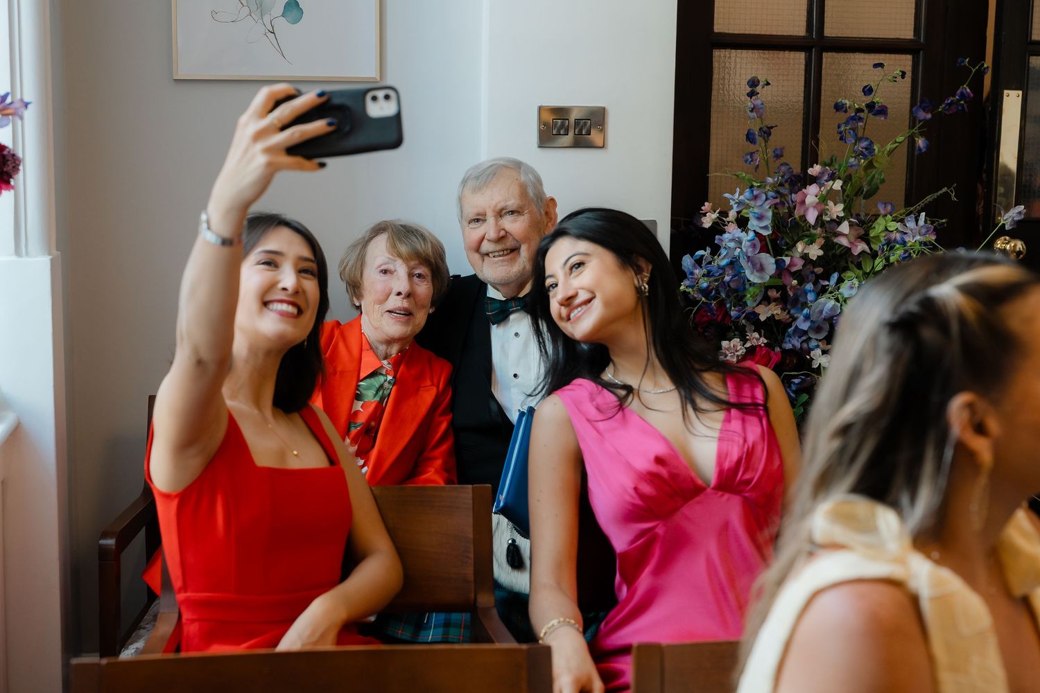 Guests taking a joyful selfie at a wedding ceremony, with an elderly couple posing with two women in red and pink dresses.