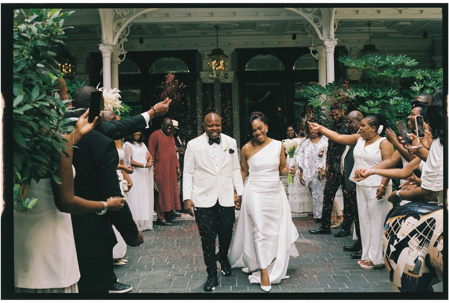 Joyful newlyweds walk through cheering guests in elegant white attire at an outdoor wedding ceremony.
