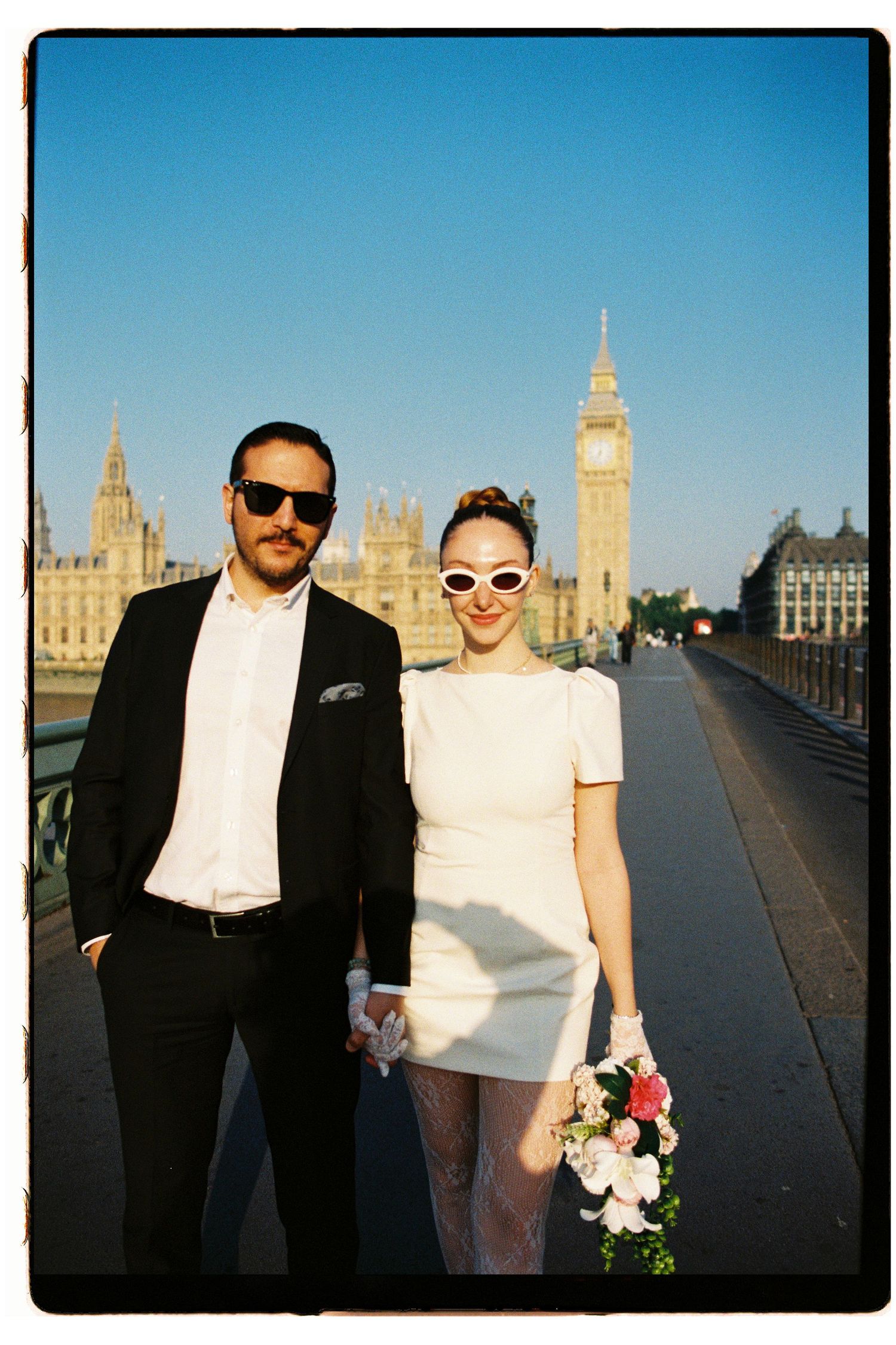 Couple in wedding attire holding hands on Westminster Bridge with Big Ben and Parliament in background.