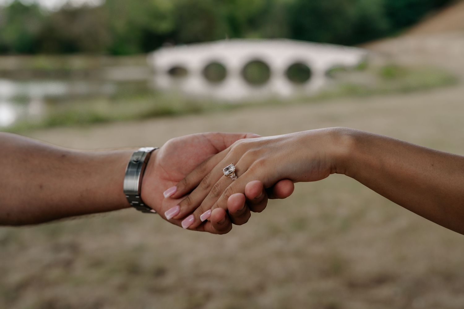 Close-up of engaged couple holding hands, woman displaying diamond engagement ring, man wearing silver watch, bridge in background.