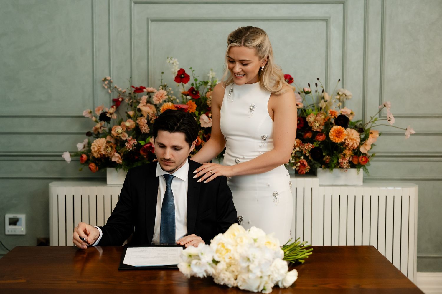 Bride stands behind groom as he signs wedding register with white bouquet nearby.