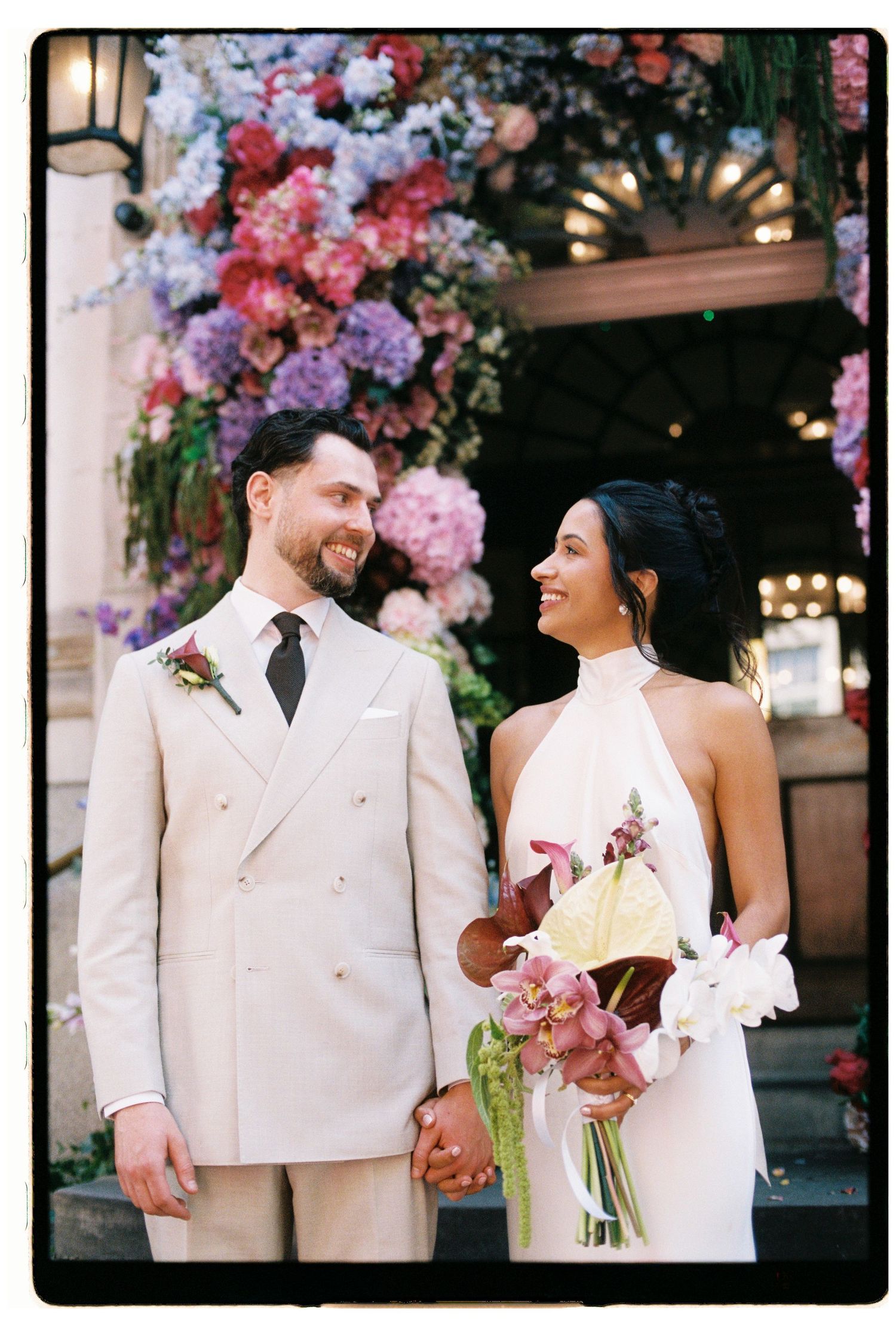 Bride and groom holding hands before stunning colorful floral wedding arch.