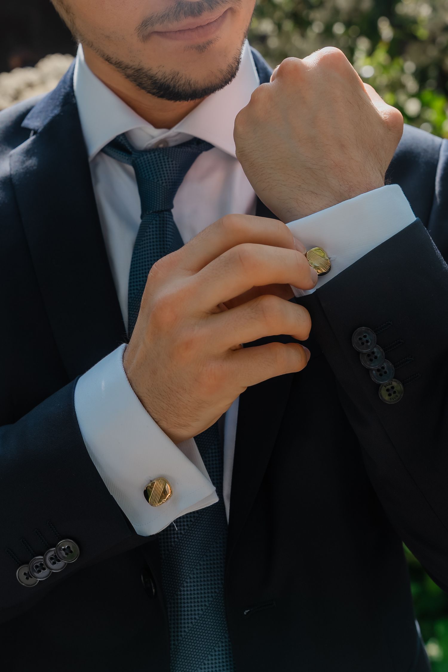 Groom in dark suit adjusts gold cufflinks while wearing teal tie on wedding day.