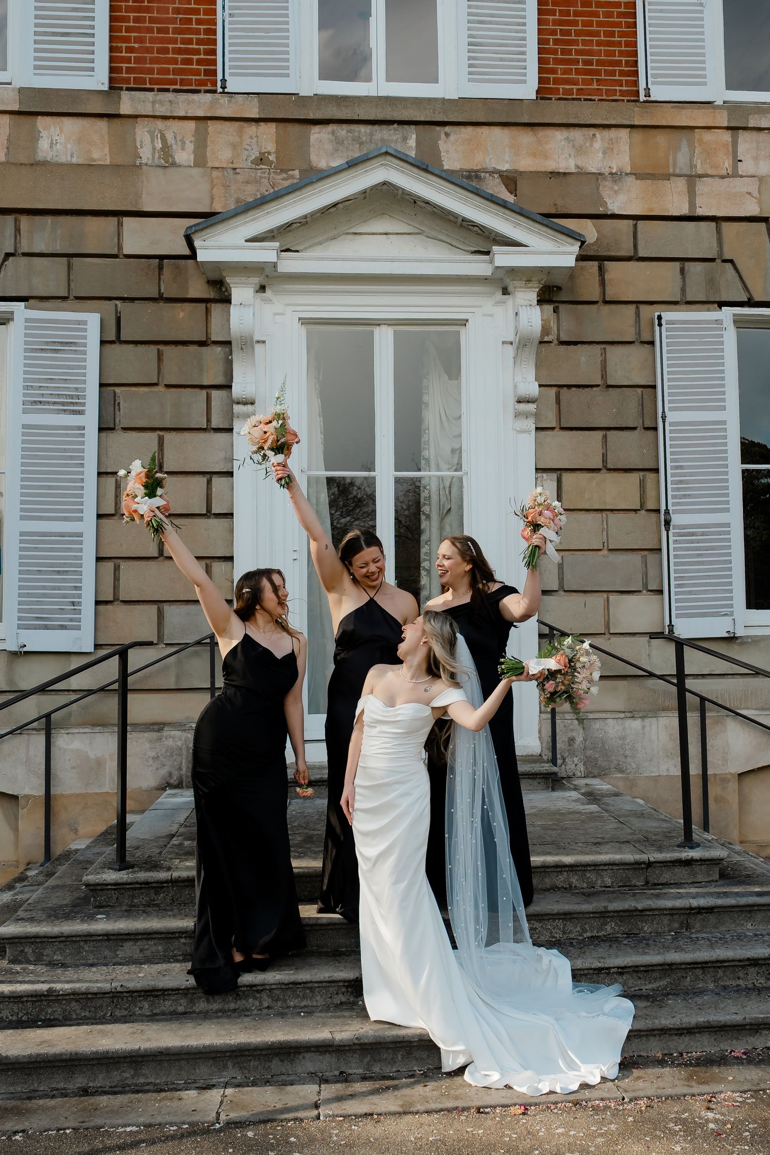 Bride poses with three bridesmaids in black dresses raising bouquets on venue steps.