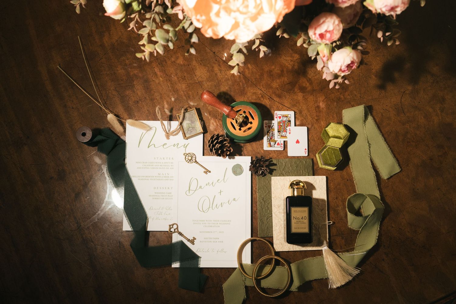 Romantic wedding flat lay with calligraphy invitation, pine cones, perfume, and sage green ribbons on wood.