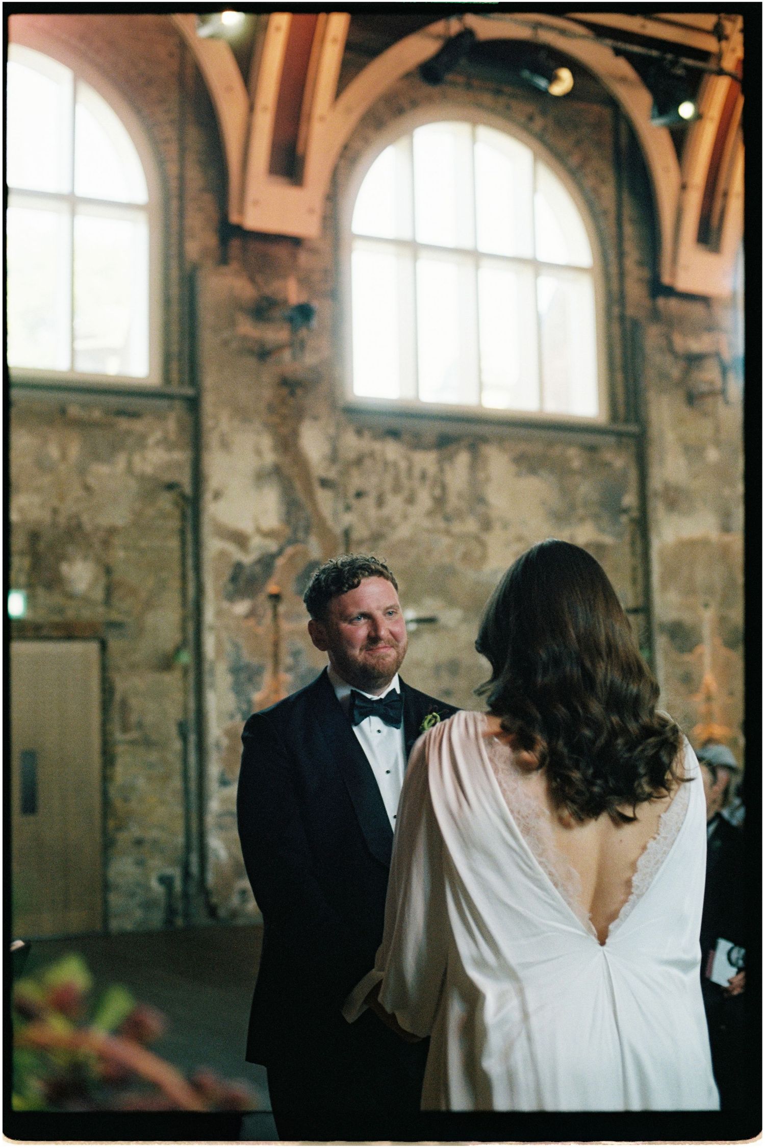Groom in black tuxedo smiles at bride during ceremony in industrial brick venue.
