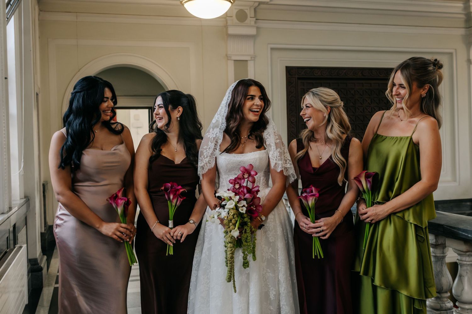 Bride in lace gown smiling with four bridesmaids in colorful satin dresses holding bouquets.