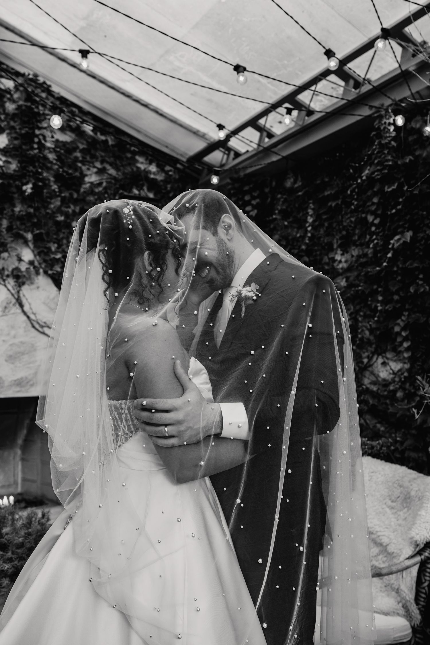 Black and white photo of bride and groom kissing beneath pearl-studded wedding veil.