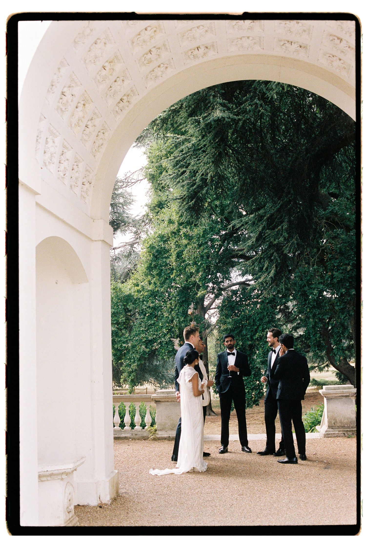 Elegant outdoor wedding ceremony under a white stone arch, with couple and officiant surrounded by lush greenery.