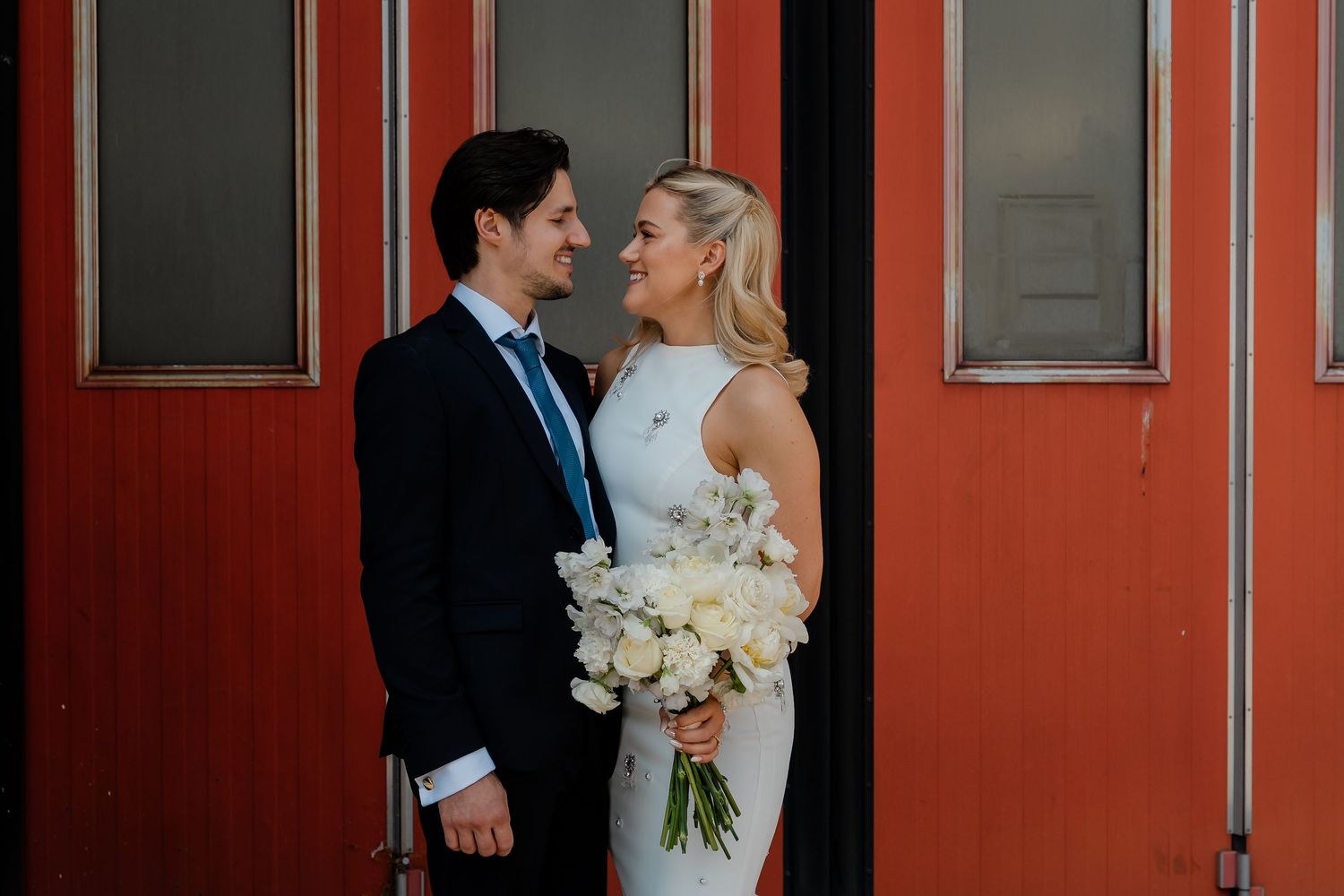 Bride and groom share a loving gaze in front of vibrant red doors holding a white floral bouquet.