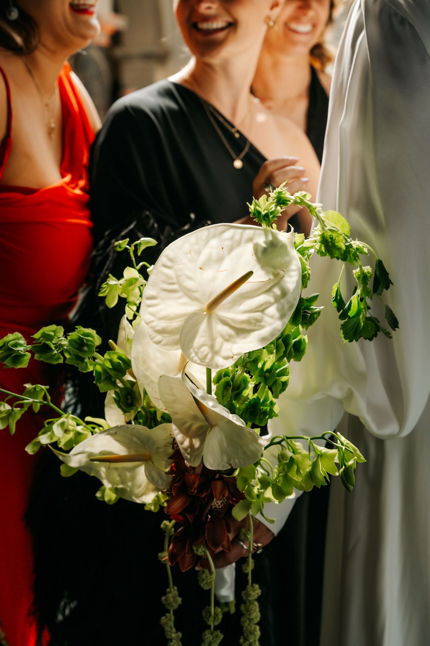 Bride holds white anthurium and green foliage bouquet surrounded by smiling guests.