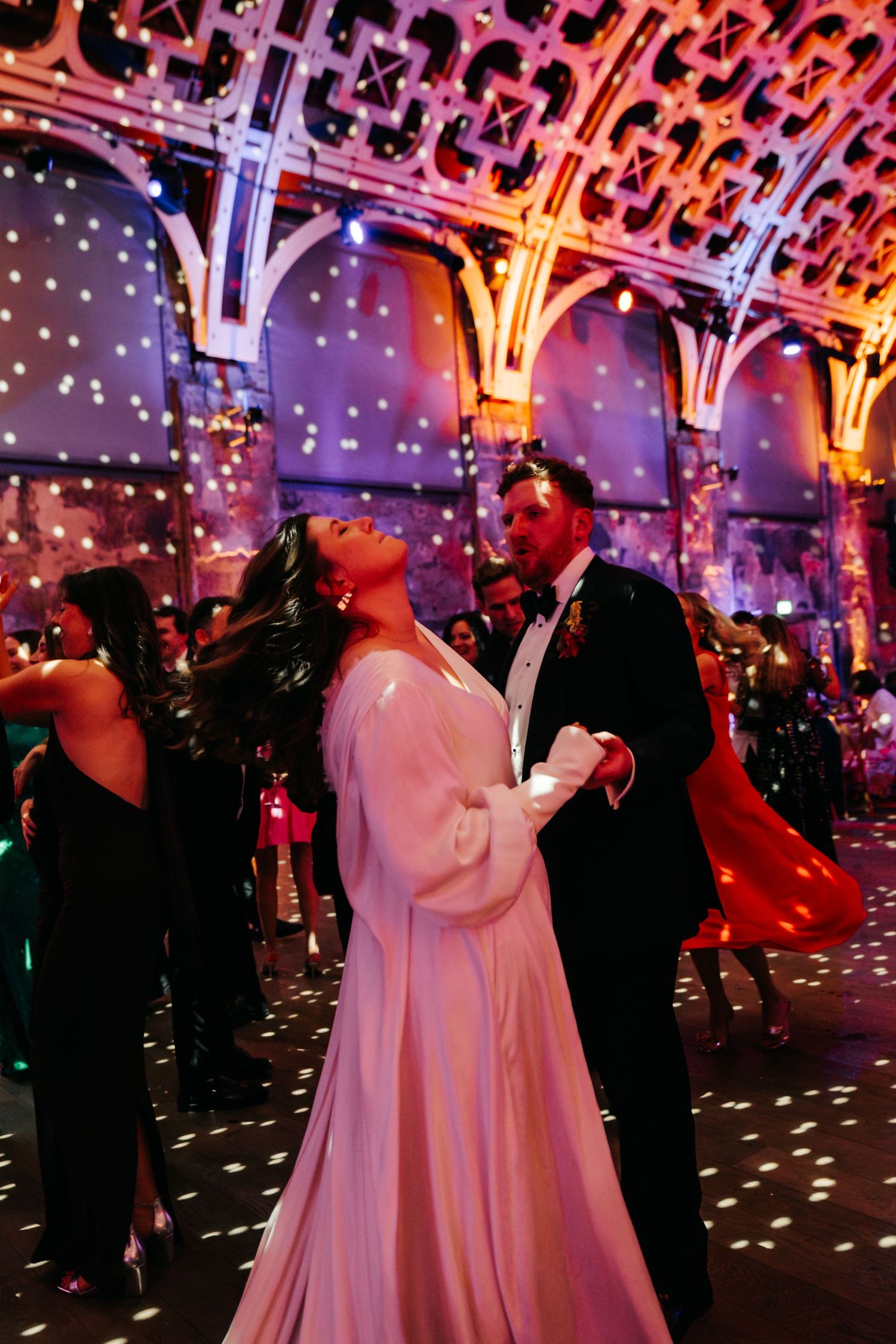 Couple dancing elegantly at a glamorous wedding reception in a grand ballroom with colorful lights and ornate ceiling.
