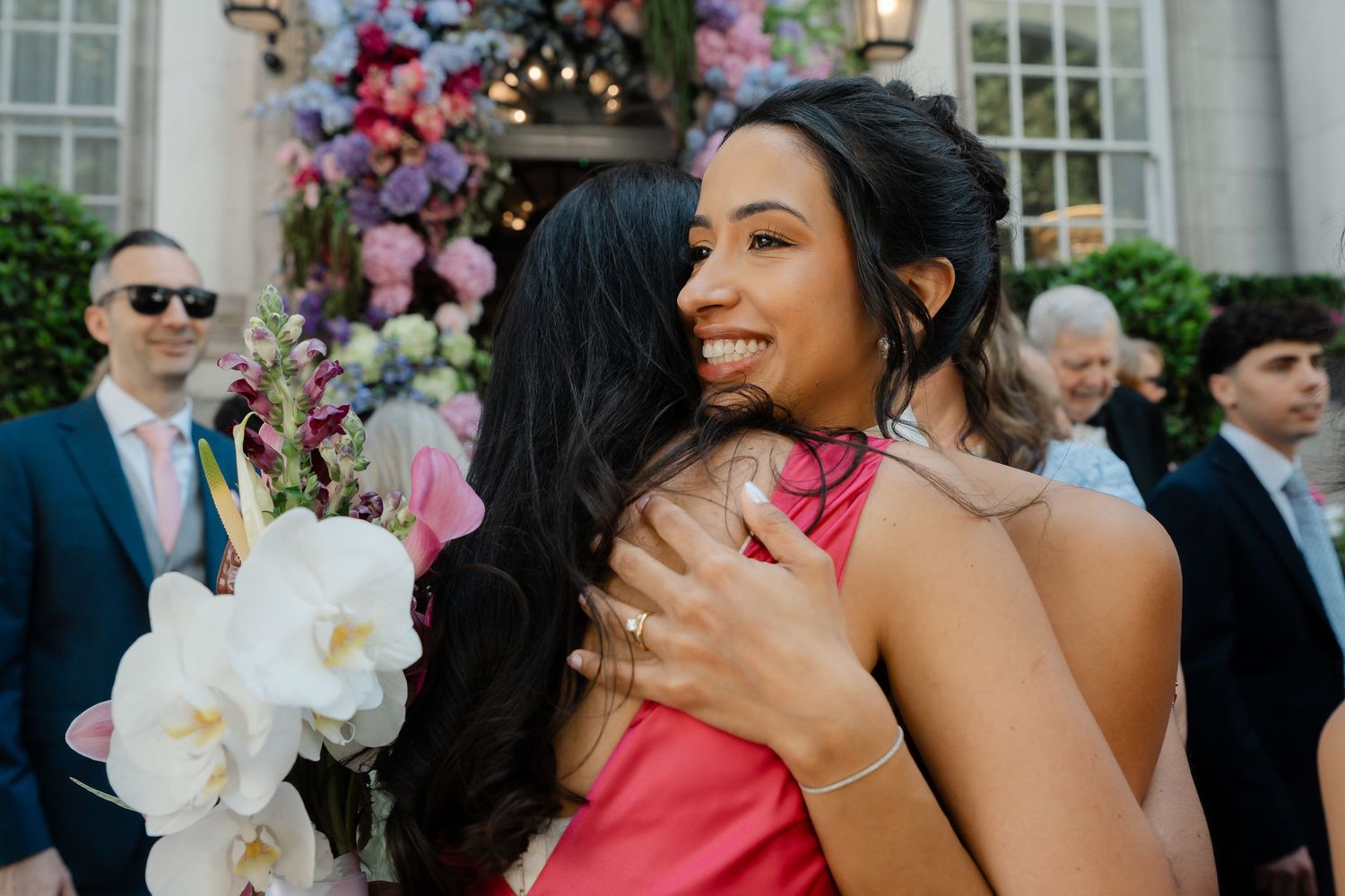 Bride in white halter dress hugging guest in pink dress at wedding.