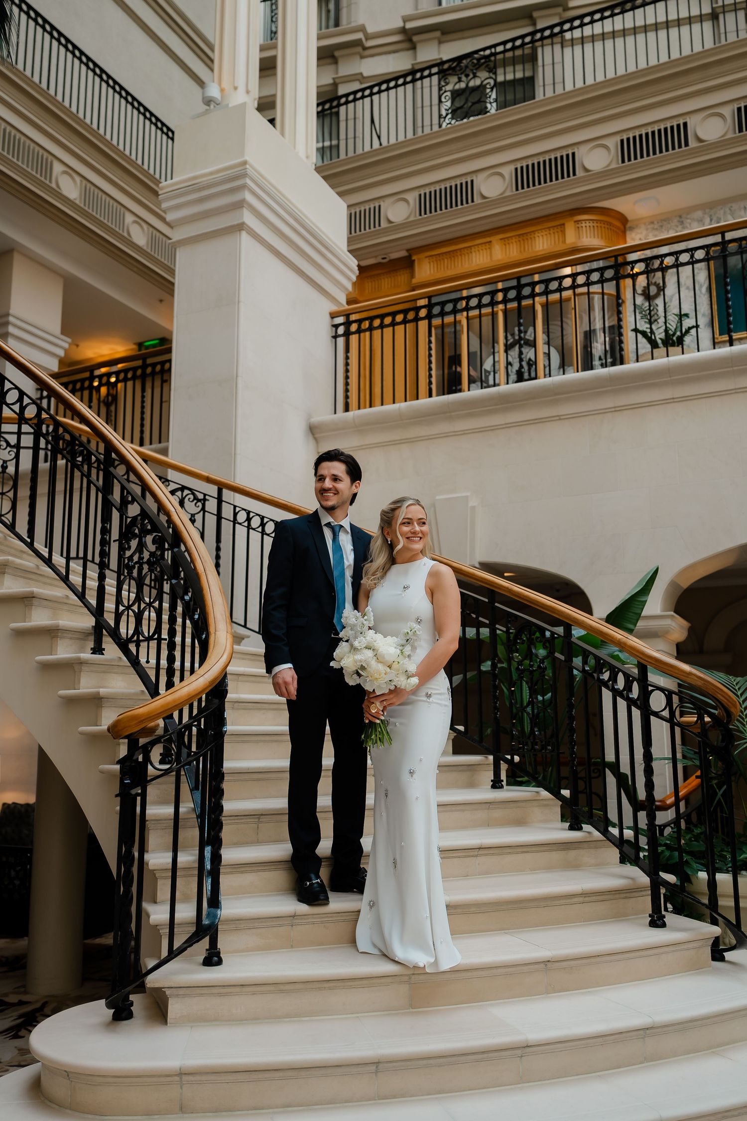 Bride and groom posing elegantly on a grand staircase with ornate railings holding a white bouquet.