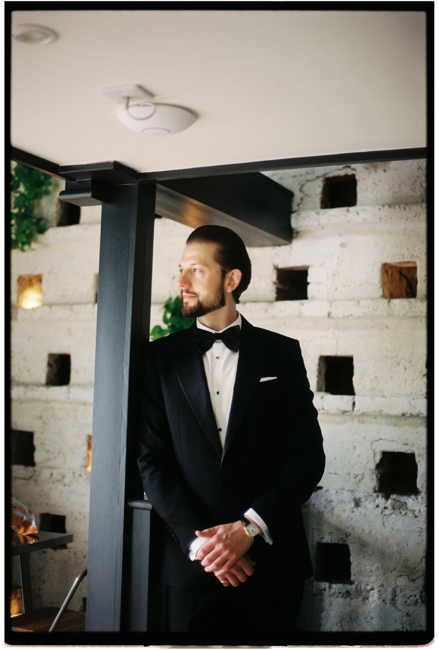 Groom in black tuxedo and bow tie stands thoughtfully beside a dark pillar in an elegant venue.