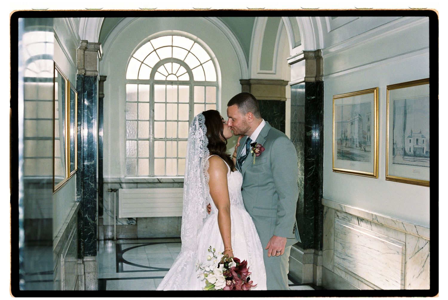 Bride and groom sharing a kiss in an elegant wedding venue hallway with arched windows and marble columns.