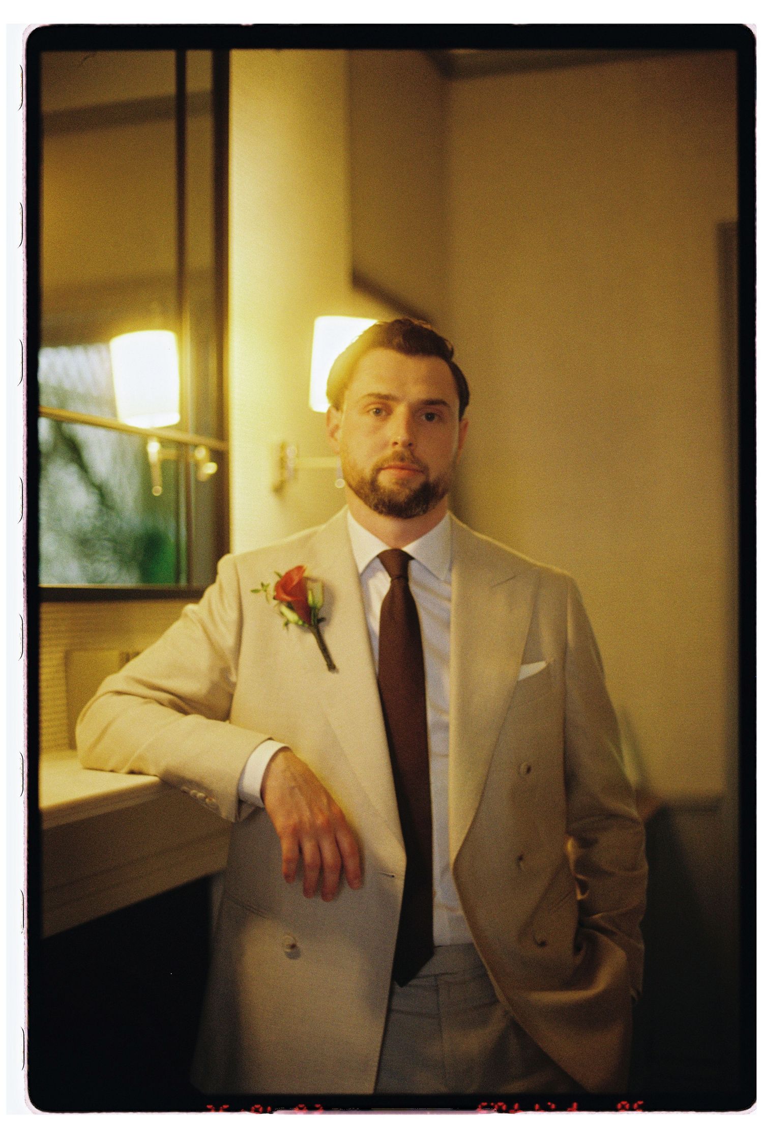 Dapper groom in beige double-breasted suit with burgundy tie and boutonniere posing indoors.