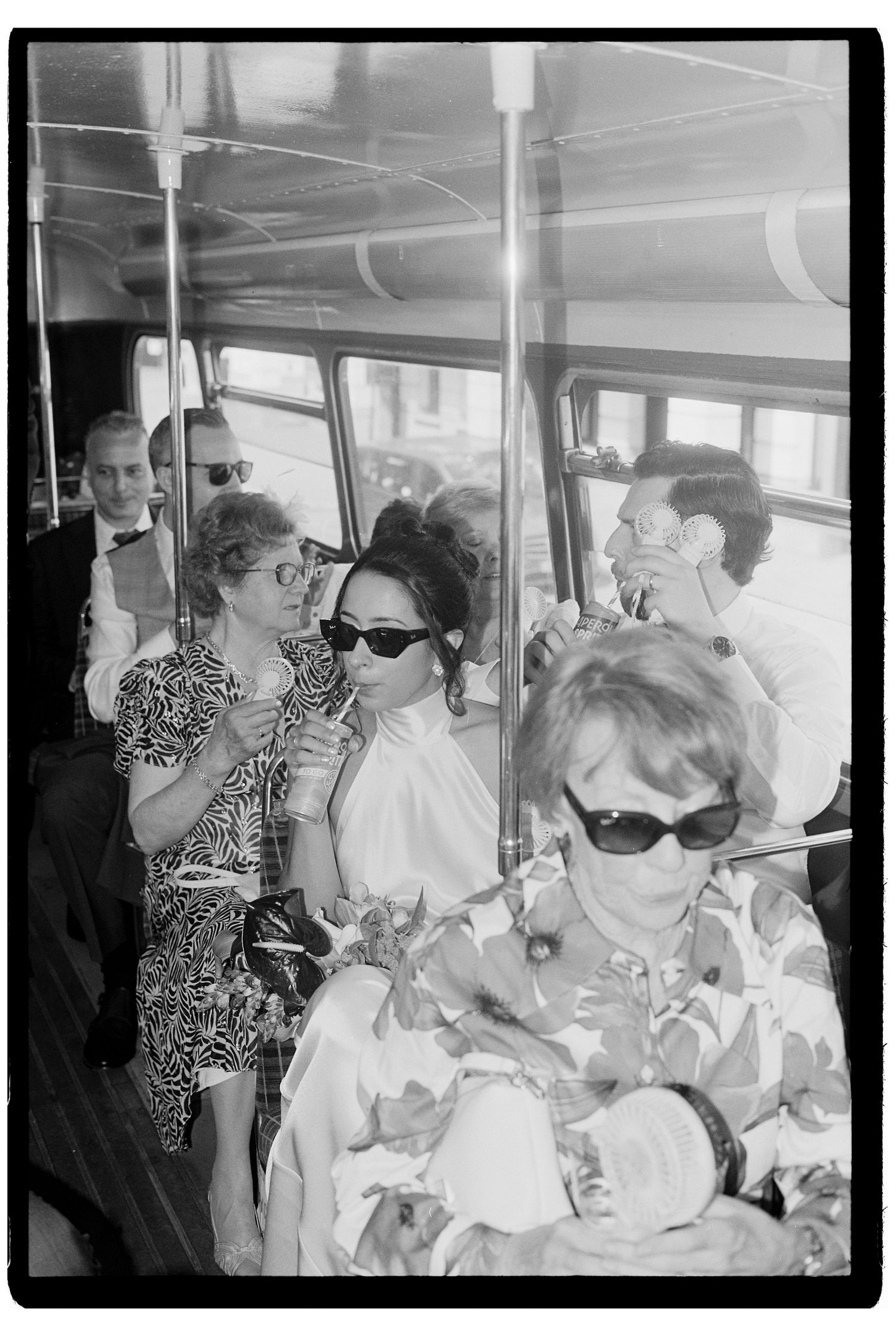 Wedding guests in sunglasses riding a vintage bus, captured in candid black and white film photography style.