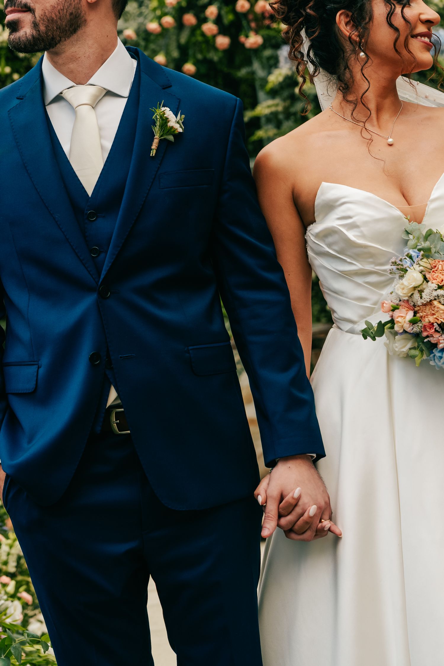 Close-up of bride and groom holding hands, groom in navy three-piece suit with boutonniere.
