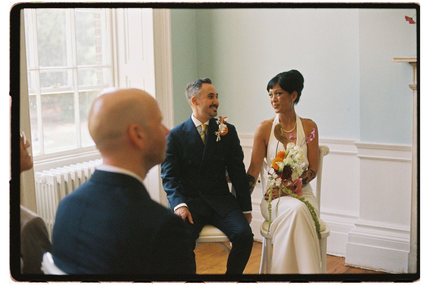 Bride and groom seated during an intimate wedding ceremony in a bright white room.