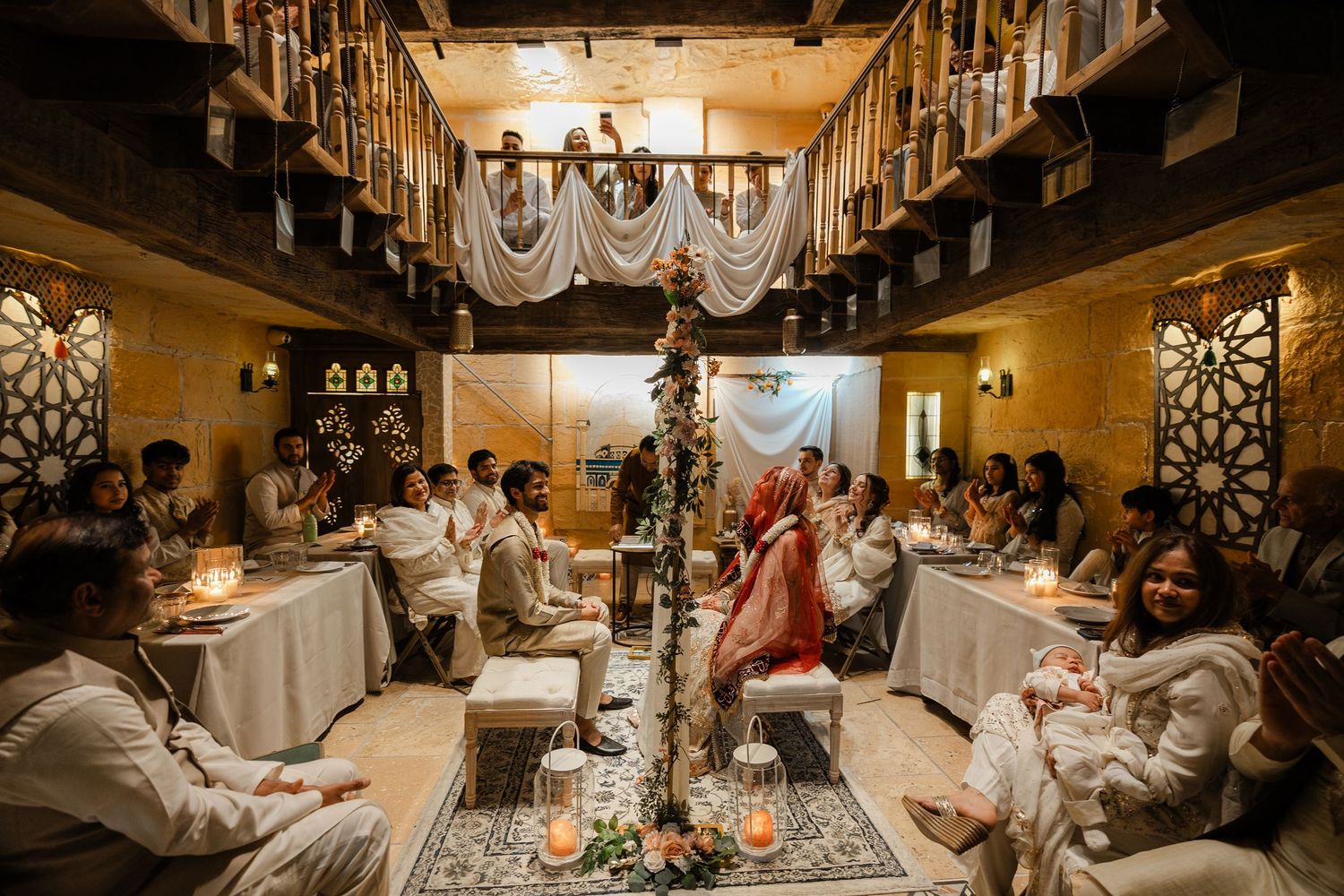 Elegant South Asian wedding ceremony in warmly lit venue with guests seated at long tables and couple at floral mandap.