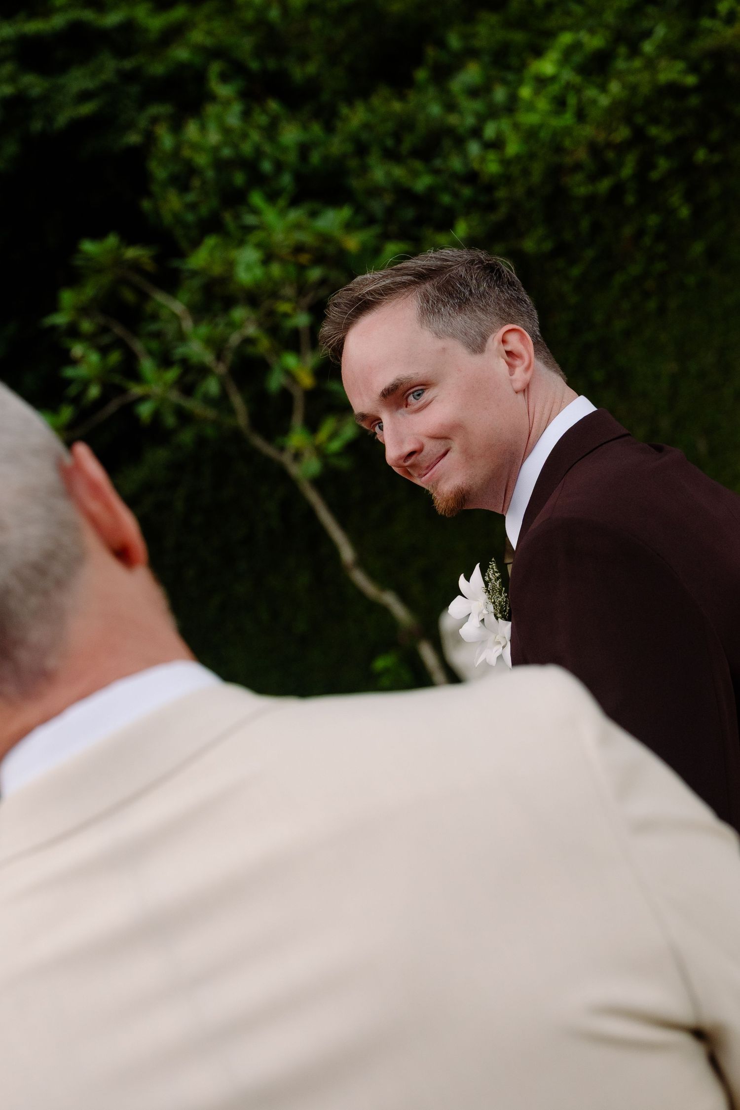 Groom in dark suit smiles warmly during outdoor wedding ceremony surrounded by lush greenery.