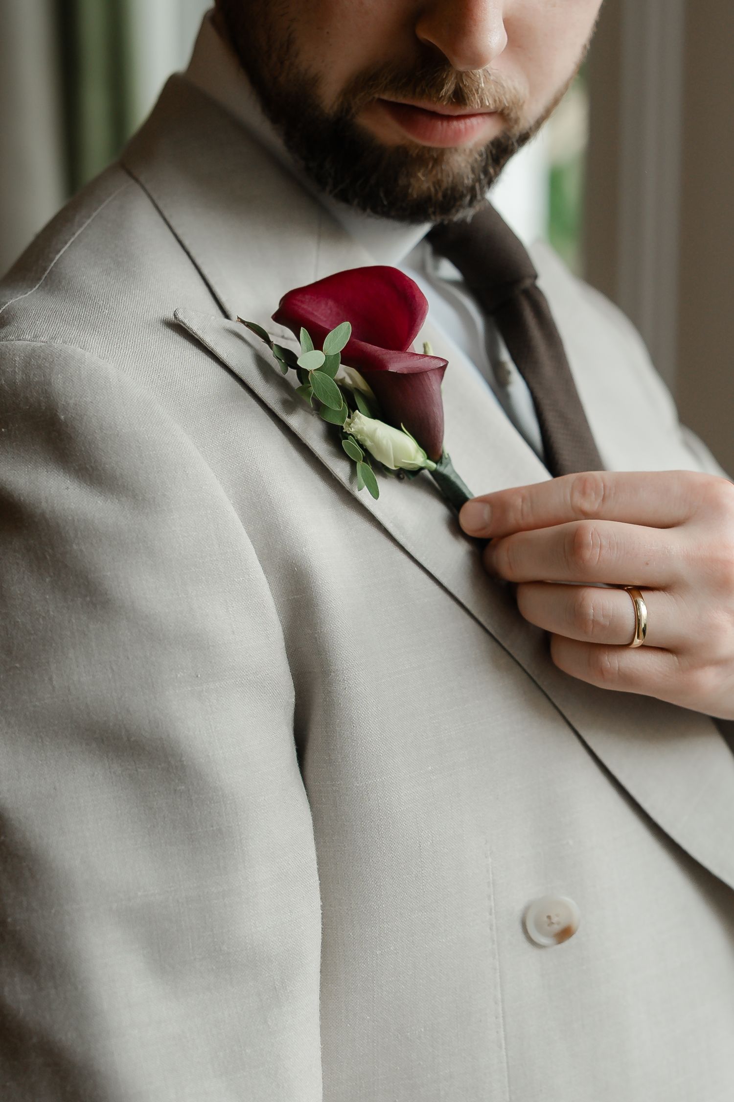Close-up of groom in beige suit with a deep red calla lily boutonniere and gold wedding band on his finger.