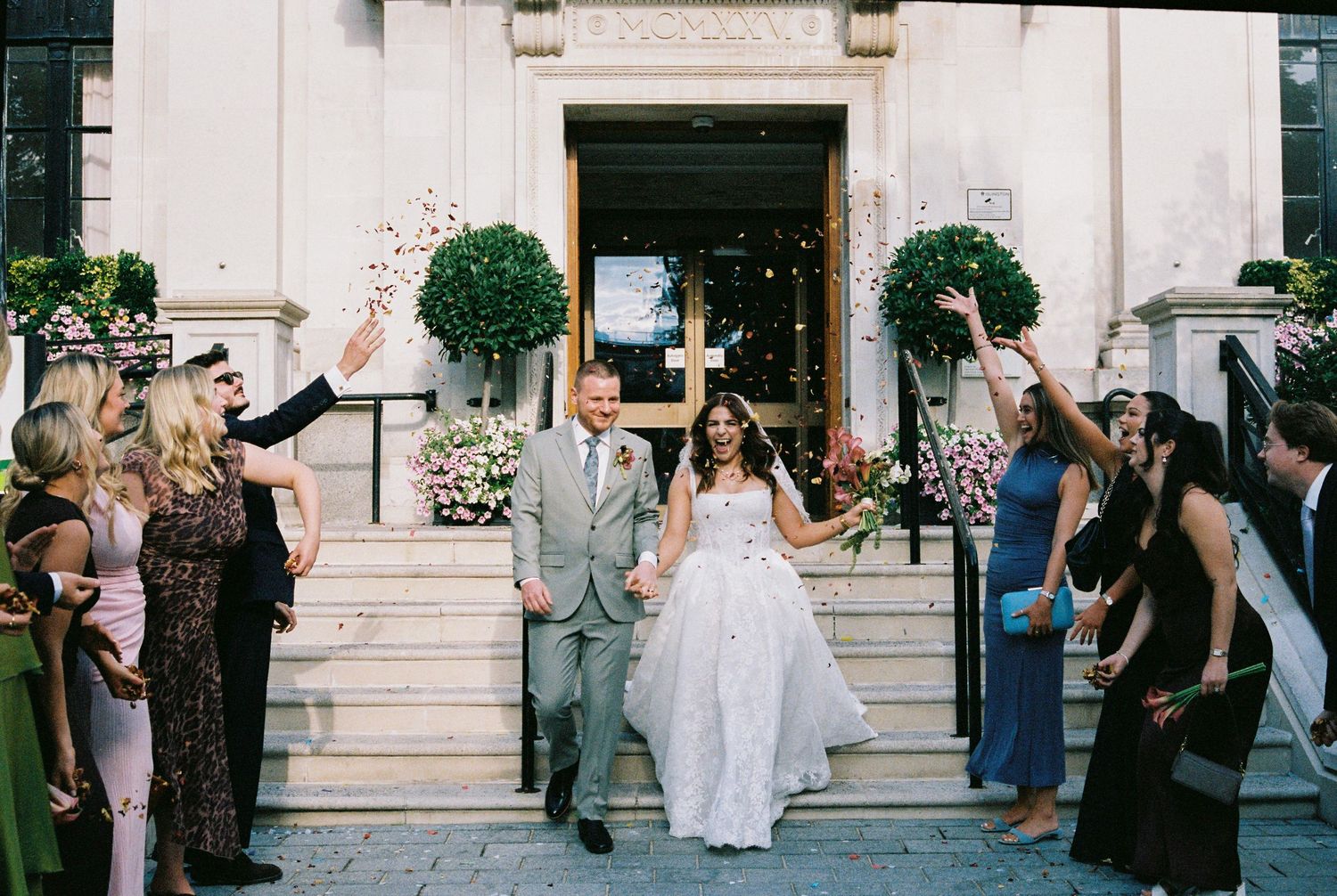 Bride in a white tulle gown and groom in a gray suit walk down church steps as cheering wedding guests celebrate on both sides.