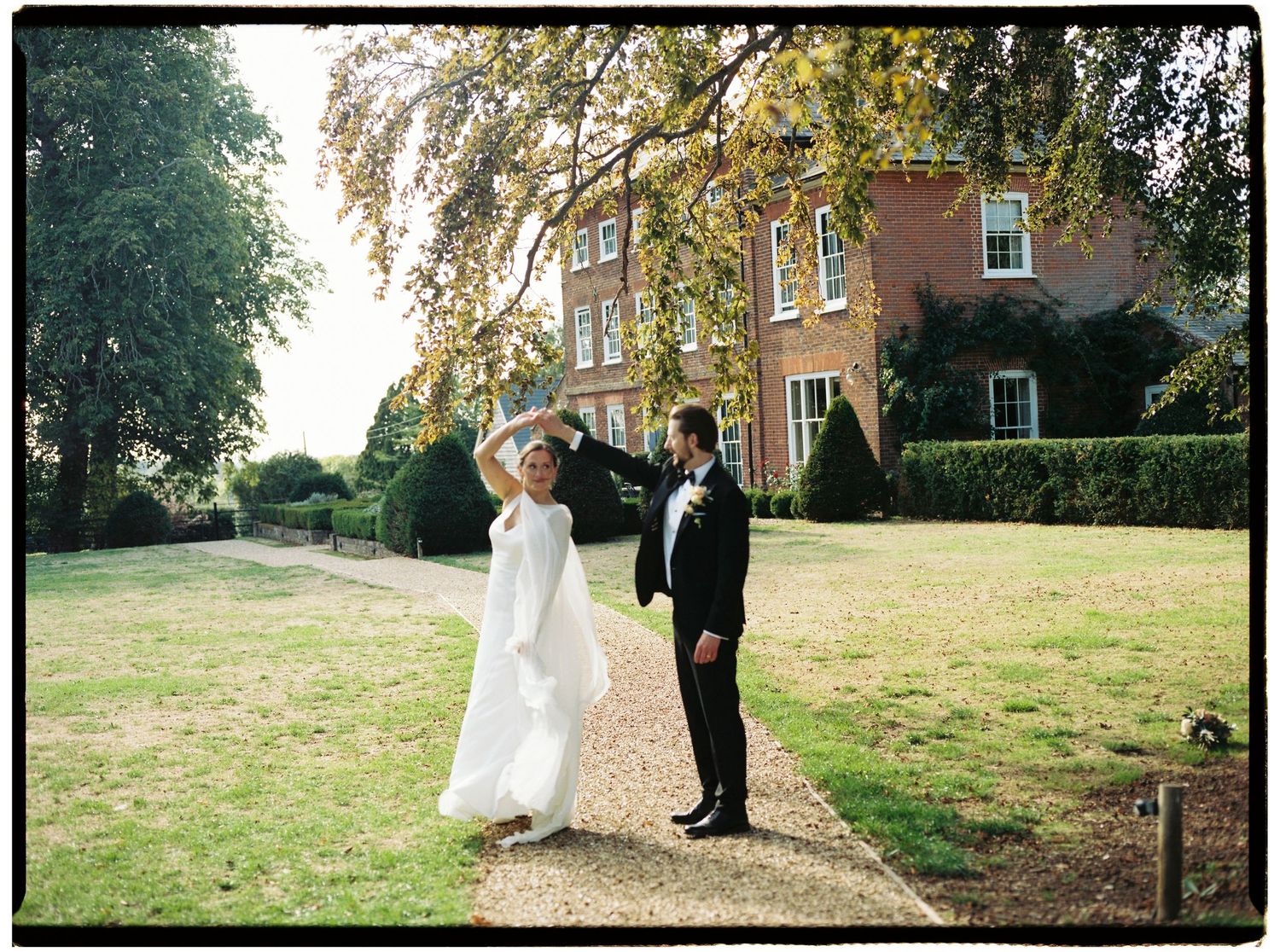 Bride and groom dancing on a garden path outside a grand brick manor house on their wedding day.