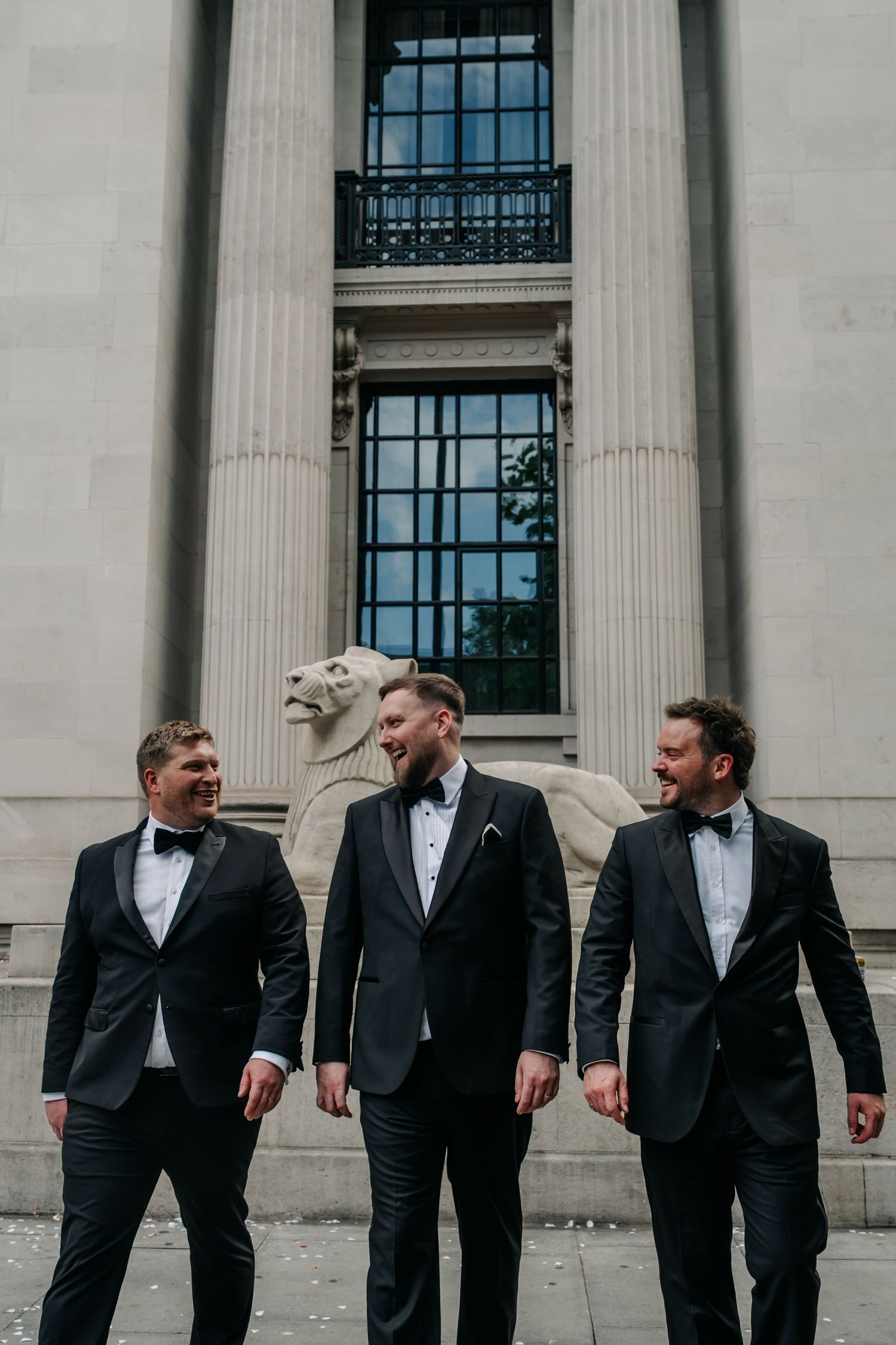Three groomsmen in black tuxedos laughing together outside grand stone building with lion sculpture.
